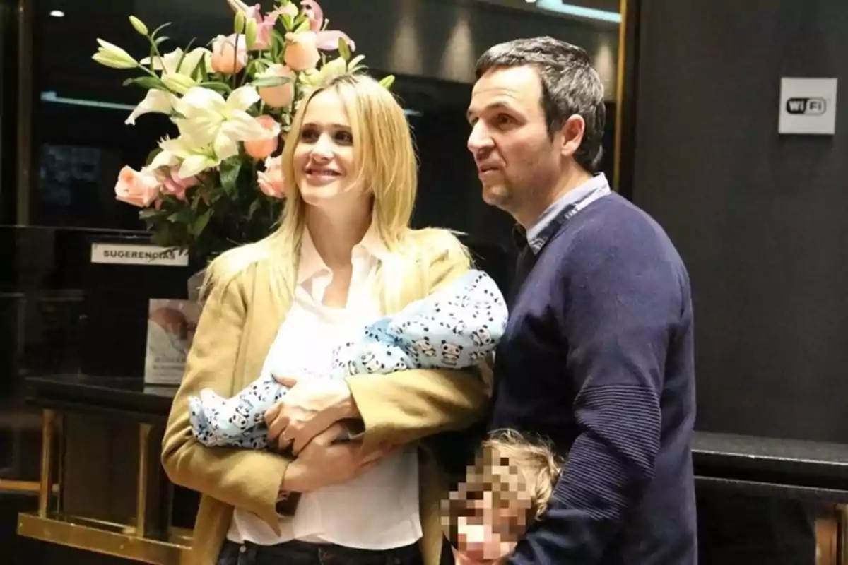 A smiling couple poses next to a pixelated toddler and a baby wrapped in a blue patterned blanket, with a bouquet of flowers in the background. A smiling couple poses next to a pixelated toddler and a baby wrapped in a blue patterned blanket, with a bouquet of flowers in the background.
