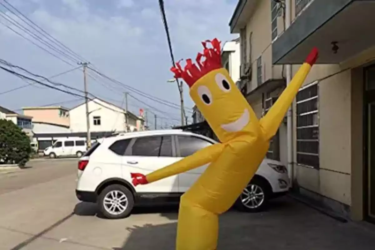 Muñeco inflable amarillo con cara sonriente y brazos extendidos frente a un edificio y un auto blanco en la calle Muñeco inflable amarillo con cara sonriente y brazos extendidos frente a un edificio y un auto blanco en la calle