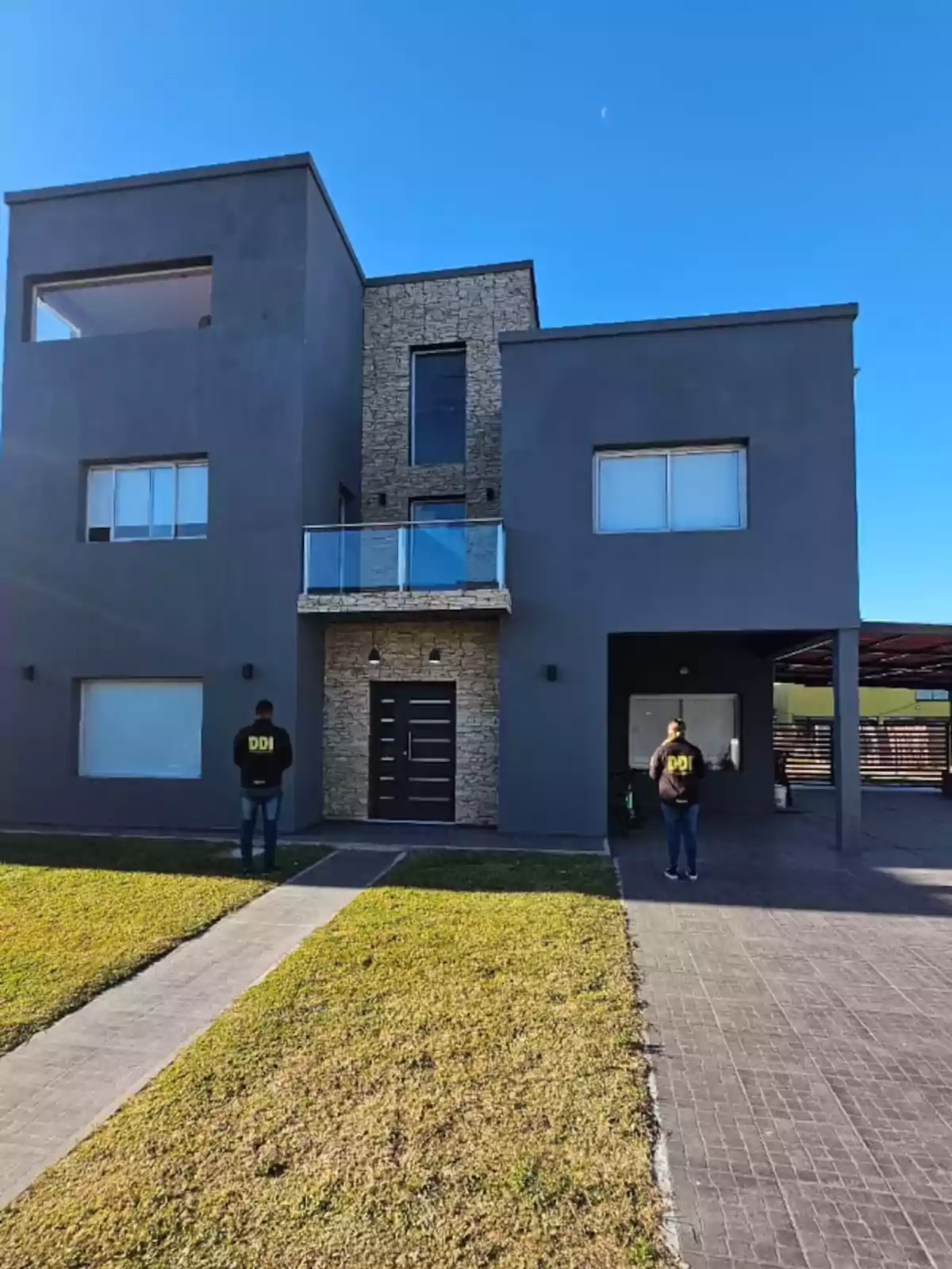 Facade of a modern two-story house with gray walls and stone details, two people wearing black jackets with the letters DDI are standing in front of the house on a sunny day. Facade of a modern two-story house with gray walls and stone details, two people wearing black jackets with the letters DDI are standing in front of the house on a sunny day.