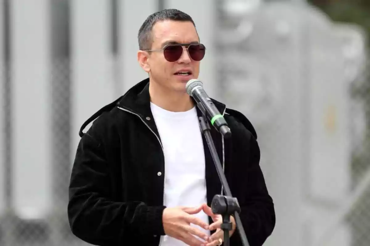 Man with sunglasses and a black jacket speaking into a microphone outdoors