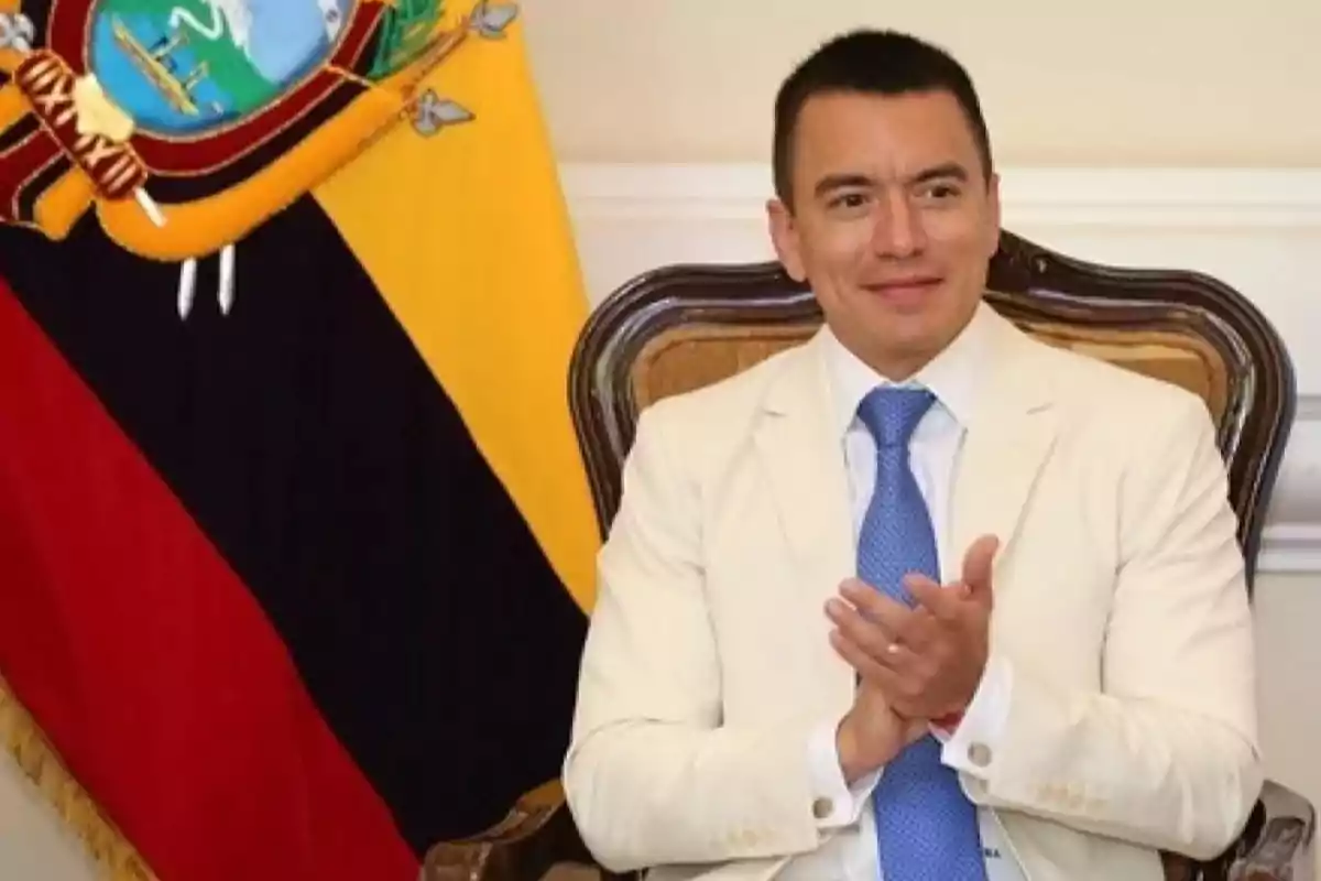 Hombre con traje claro y corbata azul sentado en una silla elegante aplaudiendo con una bandera de Ecuador al fondo Hombre con traje claro y corbata azul sentado en una silla elegante aplaudiendo con una bandera de Ecuador al fondo