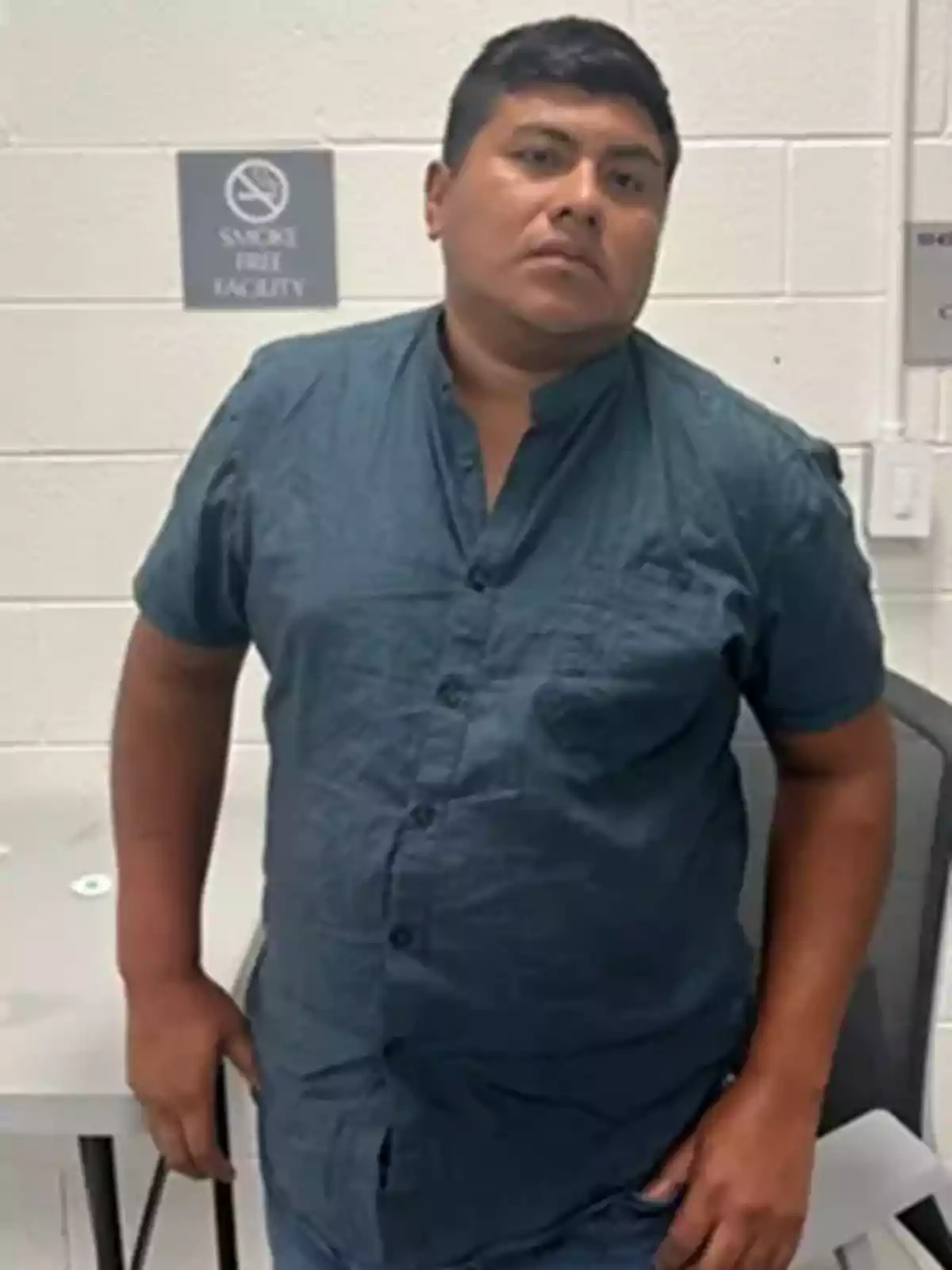 Hombre de cabello corto y camisa azul de pie junto a una mesa en una habitación con paredes blancas y señales en la pared Hombre de cabello corto y camisa azul de pie junto a una mesa en una habitación con paredes blancas y señales en la pared