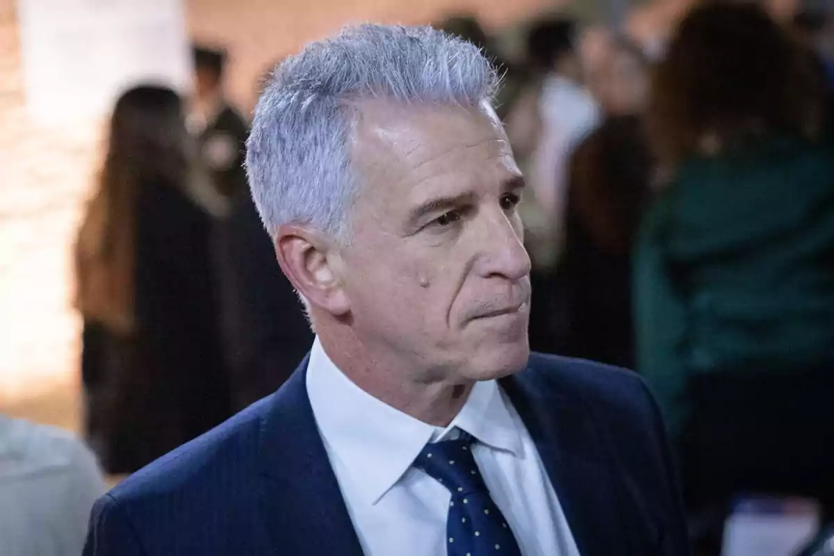 Hombre de cabello canoso con traje azul y corbata azul con puntos blancos en un evento social con personas desenfocadas al fondo