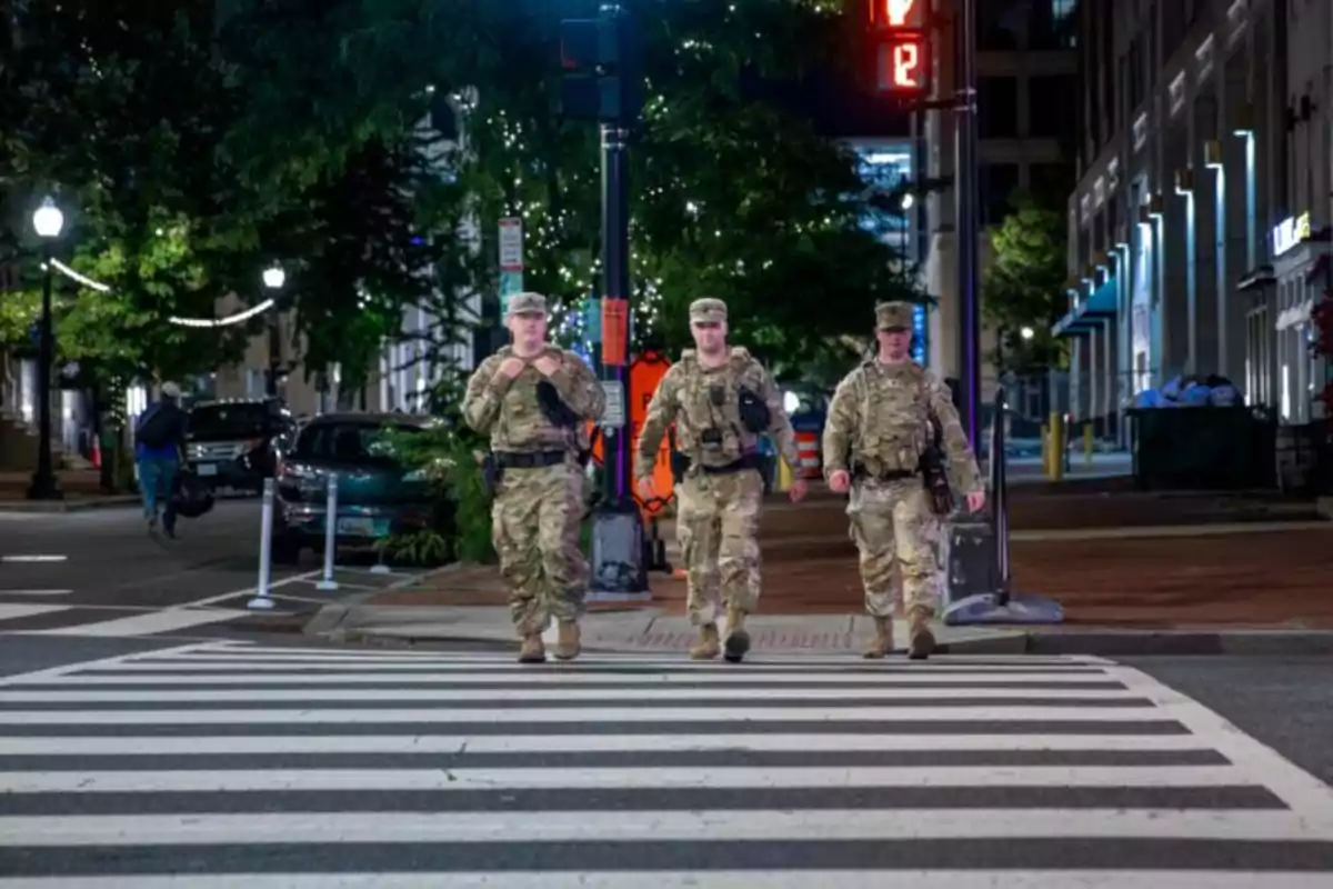 Tres soldados con uniforme de camuflaje cruzan una calle por un paso de cebra en una ciudad iluminada por la noche Tres soldados con uniforme de camuflaje cruzan una calle por un paso de cebra en una ciudad iluminada por la noche