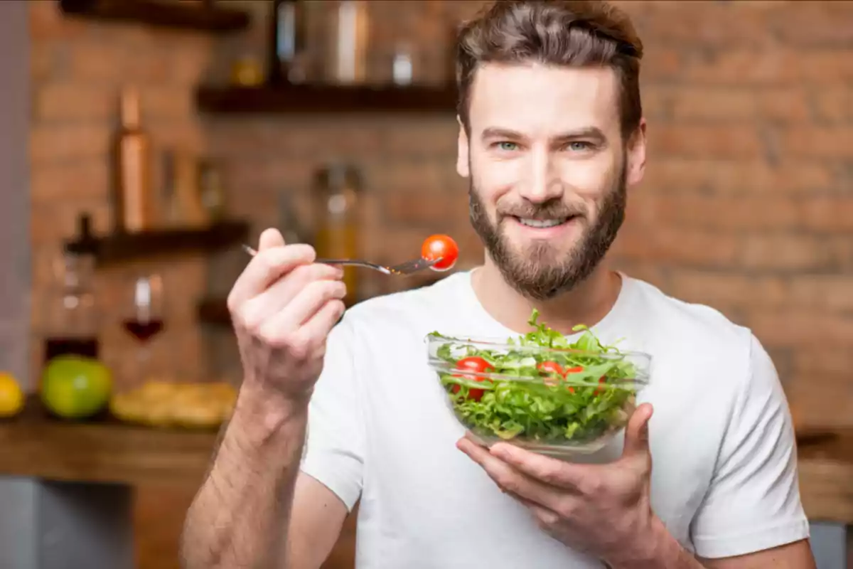 Smiling man holds a bowl of salad and a fork with a cherry tomato in a kitchen Smiling man holds a bowl of salad and a fork with a cherry tomato in a kitchen