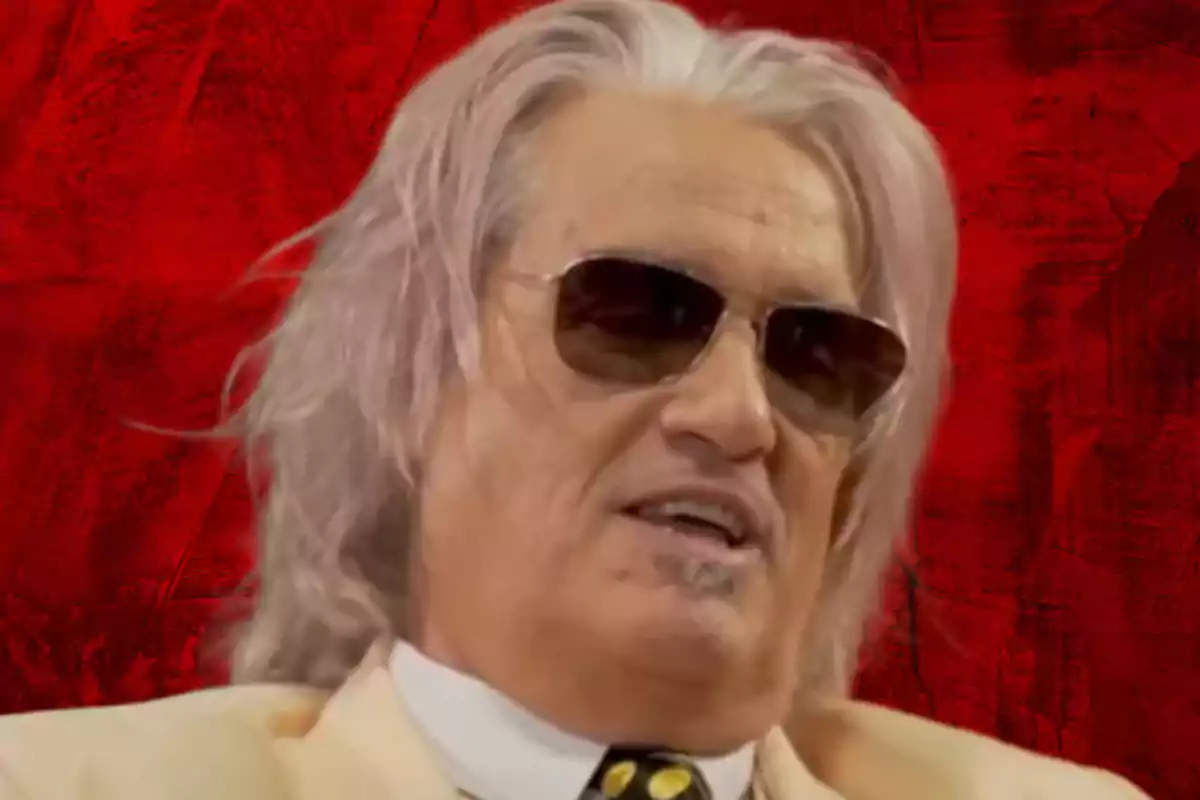 Gray-haired, long-haired man with sunglasses and a light suit against a red background