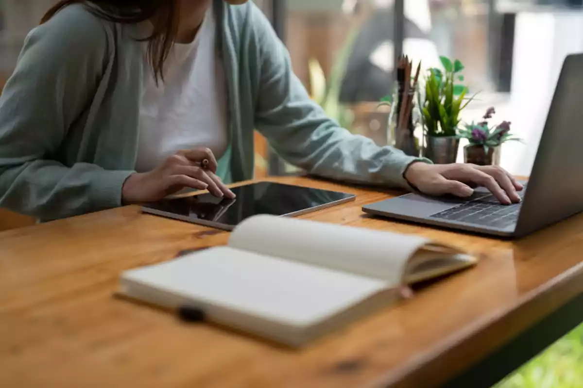 Person working at a wooden desk with a laptop, a tablet, and an open notebook, surrounded by plants and office supplies Person working at a wooden desk with a laptop, a tablet, and an open notebook, surrounded by plants and office supplies
