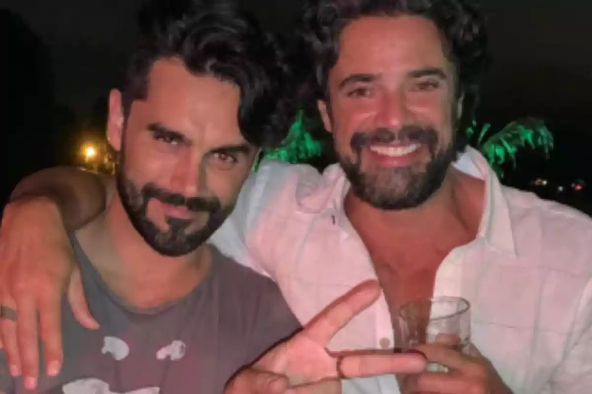 Two men smile and pose together in a nighttime photo, one of them holding a glass and making the peace sign with his fingers. Two men smile and pose together in a nighttime photo, one of them holding a glass and making the peace sign with his fingers.