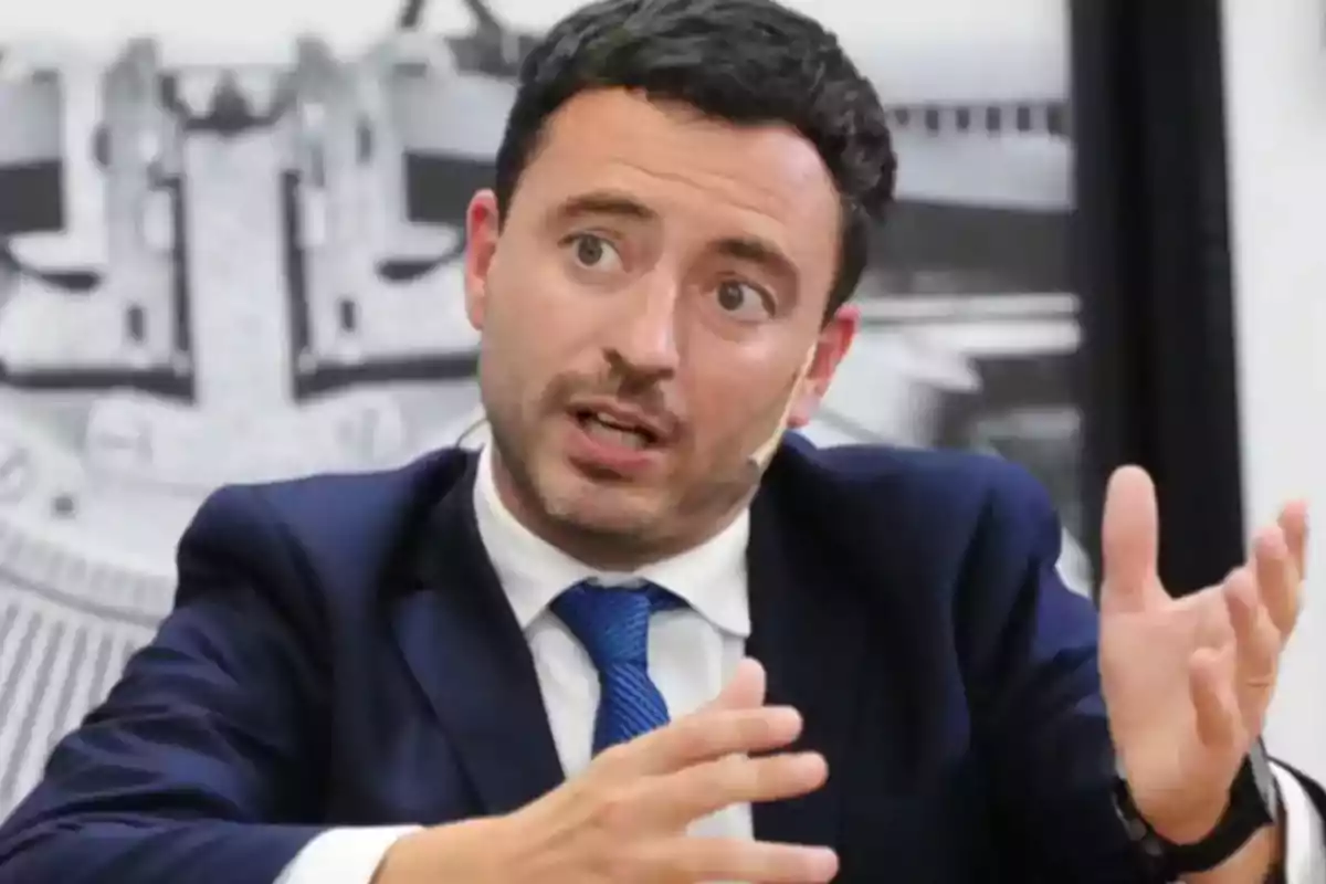 Hombre de cabello corto y oscuro con traje azul y corbata azul hablando y gesticulando con las manos durante una entrevista o conferencia Hombre de cabello corto y oscuro con traje azul y corbata azul hablando y gesticulando con las manos durante una entrevista o conferencia