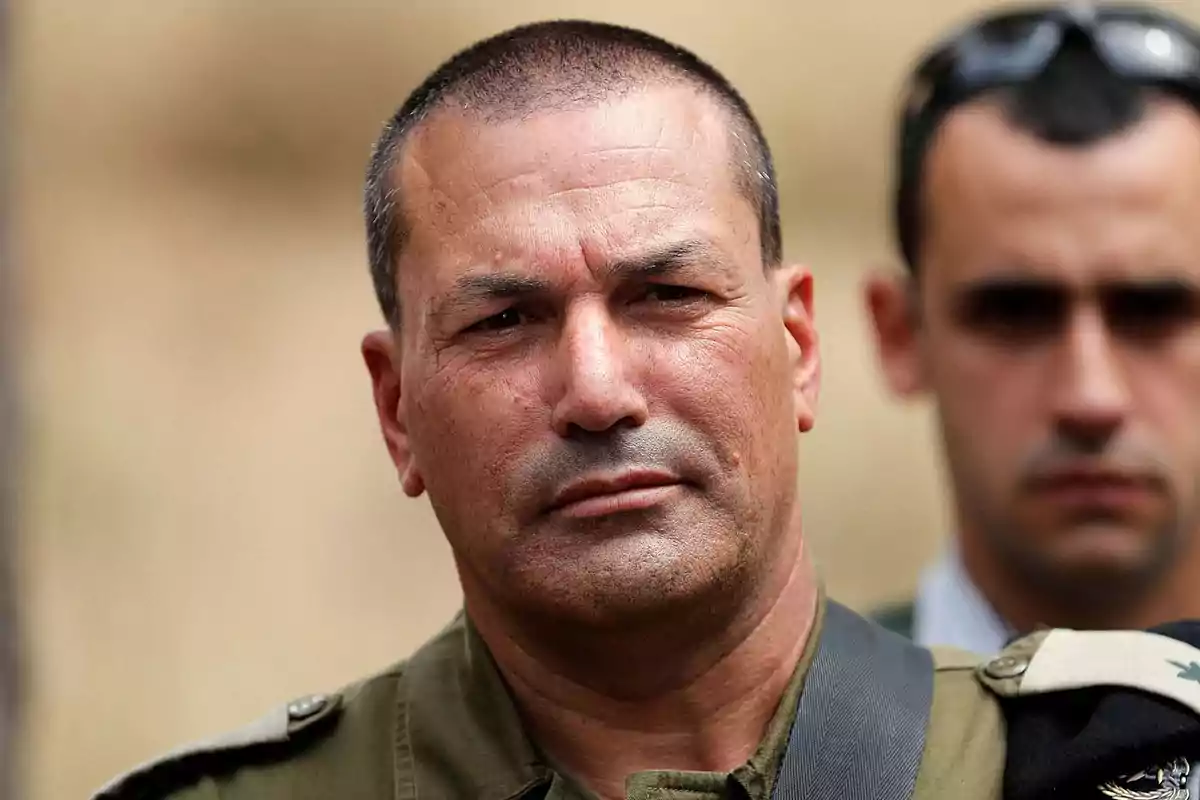 Hombre de cabello corto con uniforme militar mirando al frente con expresión seria y otra persona desenfocada al fondo