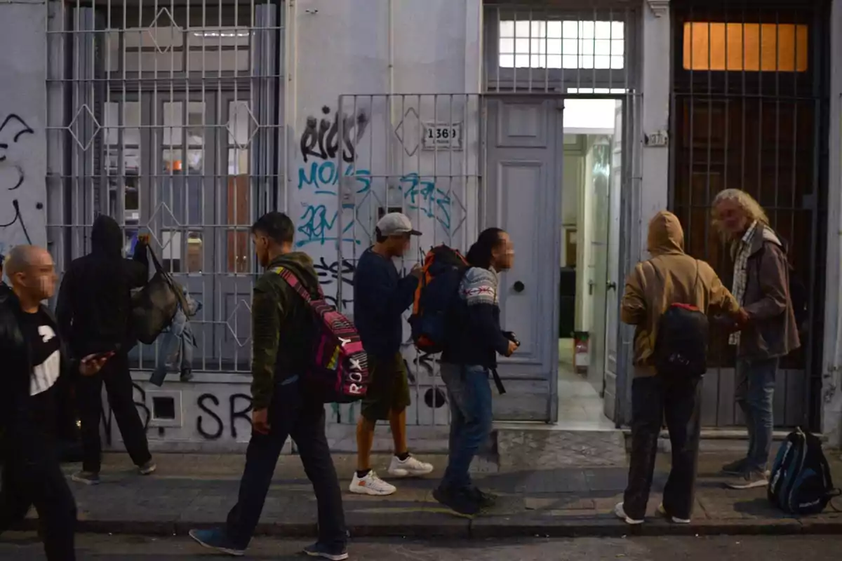 Several people with backpacks and casual clothing are gathered in front of the entrance of a building on an urban street with graffiti-covered walls.