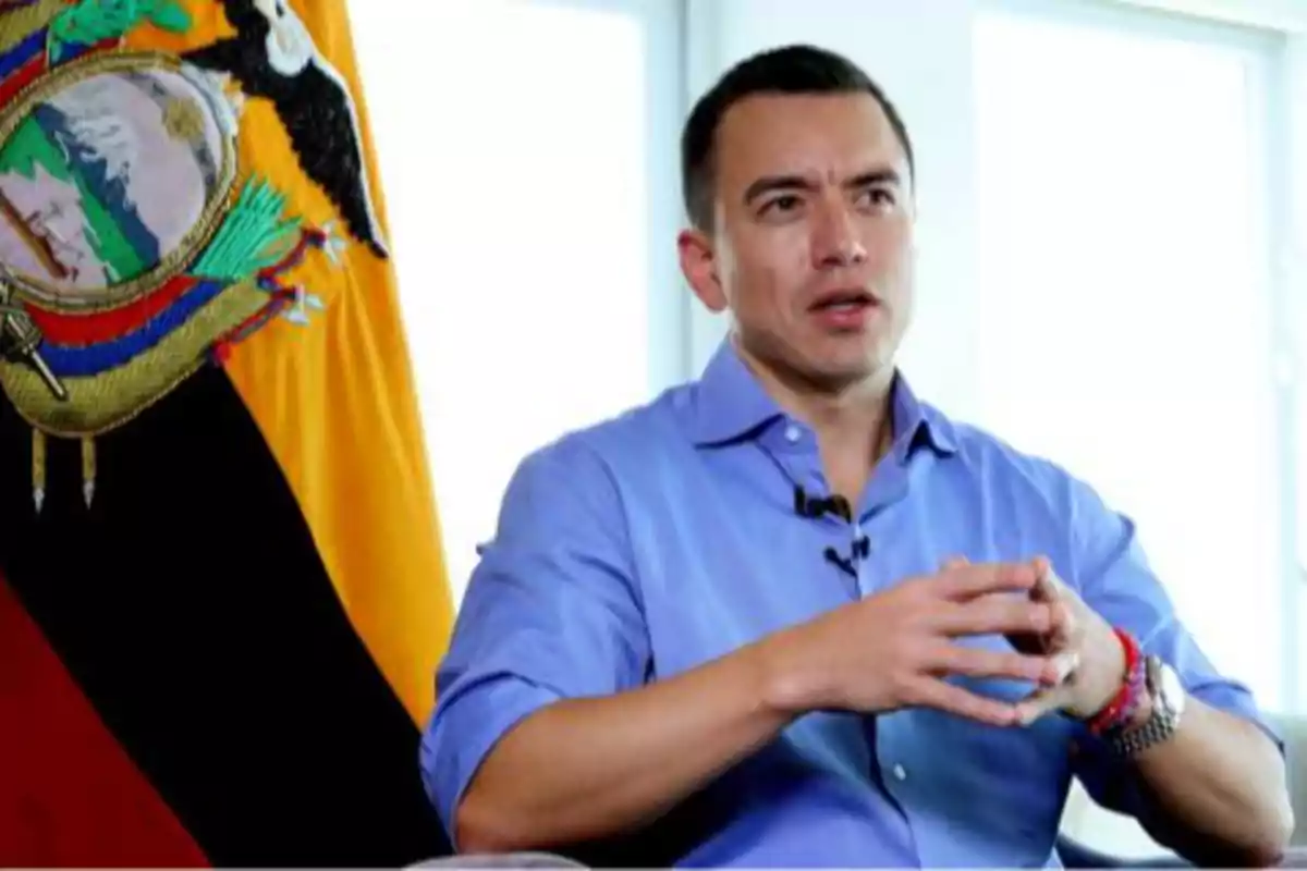 Hombre con camisa azul sentado frente a una bandera de Ecuador mientras habla y gesticula con las manos