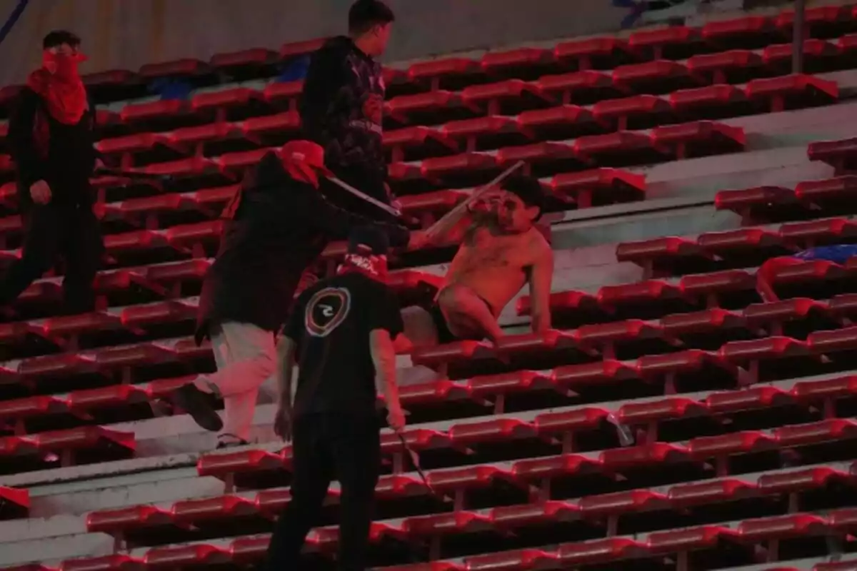 Un grupo de personas con el rostro cubierto agrede a un hombre sin camisa que está sentado en las gradas de un estadio.