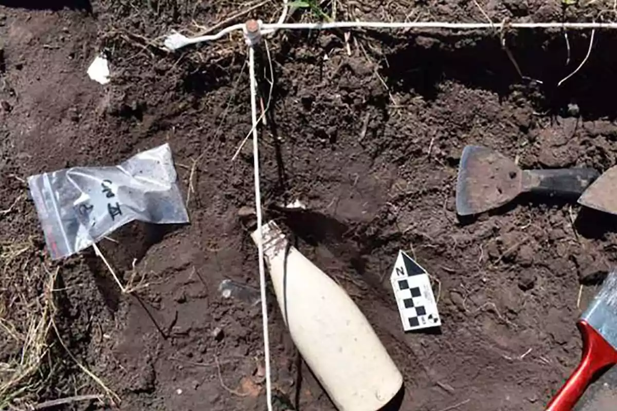Botella antigua parcialmente enterrada en tierra junto a herramientas de excavación, una bolsa de plástico con evidencia y una escala de medición arqueológica Botella antigua parcialmente enterrada en tierra junto a herramientas de excavación, una bolsa de plástico con evidencia y una escala de medición arqueológica