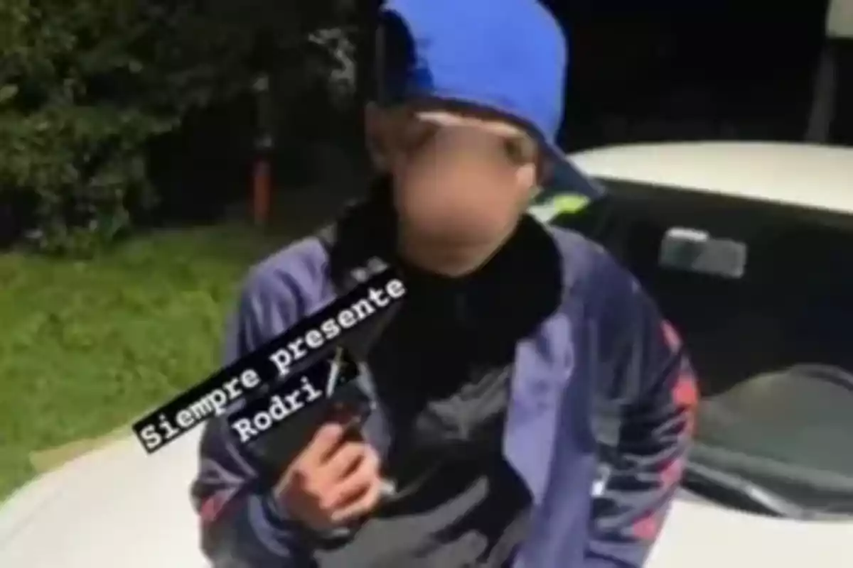 Young man with a blue cap worn backwards and a blurred face posing in front of a white car, holding an object in his hand and an overlaid text that says Always present Rodri