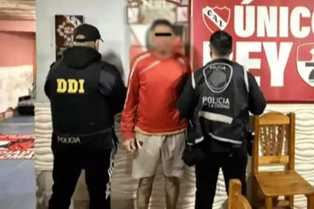 Dos policías detienen a un hombre con el rostro difuminado frente a un cartel rojo en una habitación