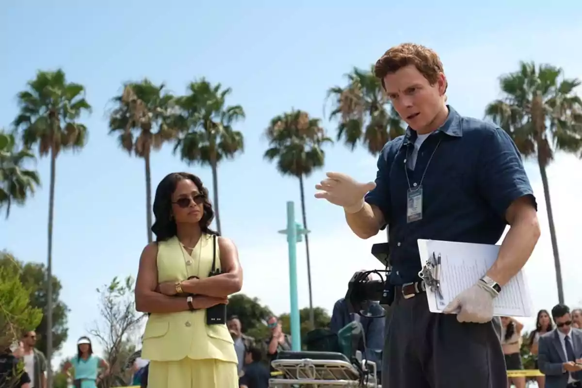 Two people are talking outdoors surrounded by palm trees; one of them is wearing gloves and holding a folder while the other watches with their arms crossed. Two people are talking outdoors surrounded by palm trees; one of them is wearing gloves and holding a folder while the other watches with their arms crossed.
