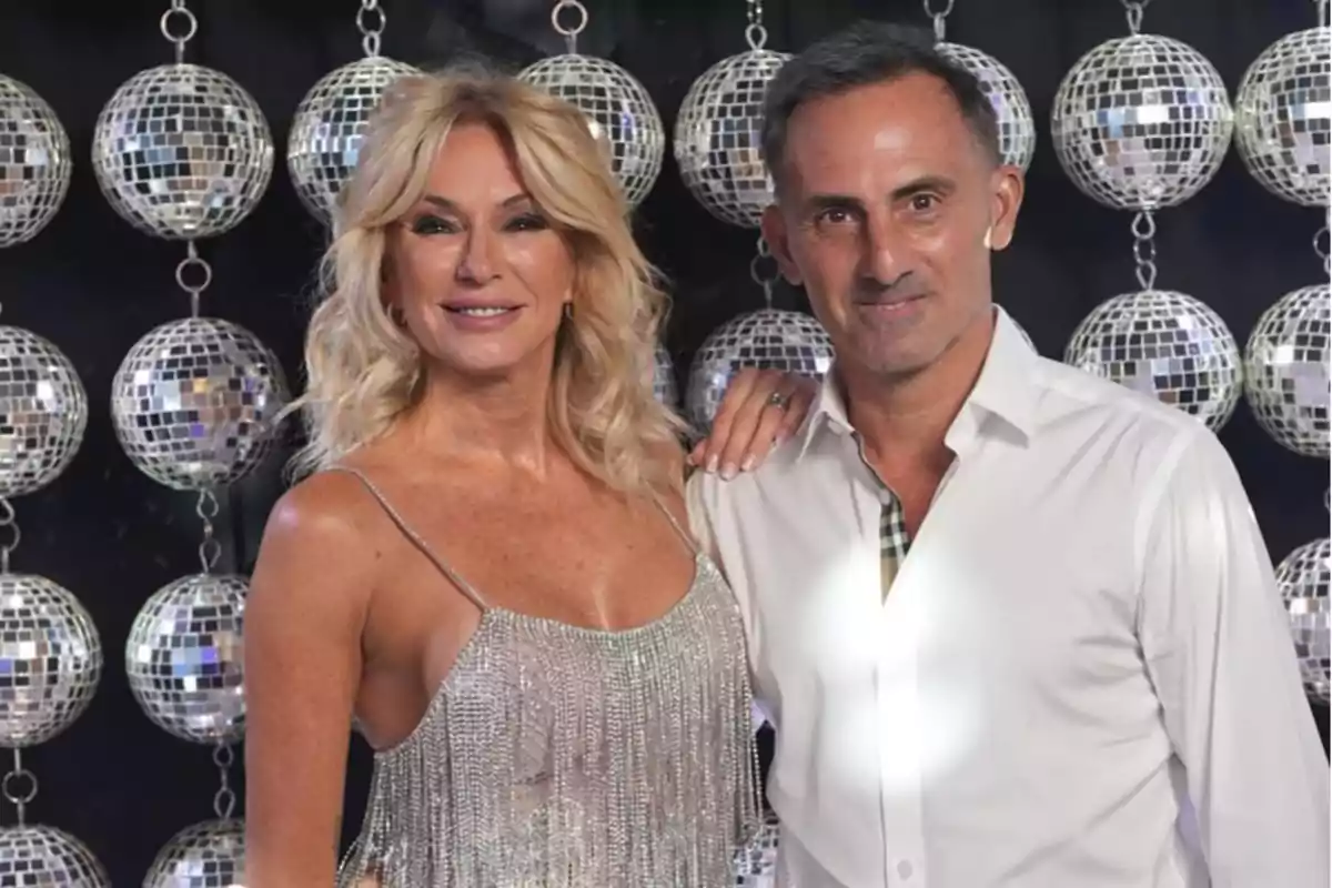Una mujer rubia con vestido plateado y un hombre de camisa blanca posan juntos frente a un fondo de esferas de espejos brillantes