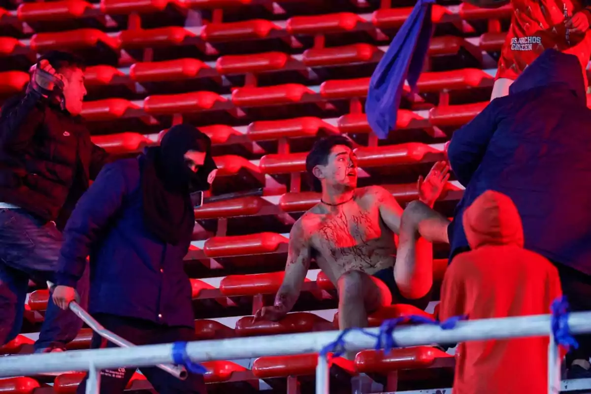 Un hombre herido y cubierto de sangre está sentado en las gradas de un estadio mientras varias personas lo rodean, algunas con el rostro cubierto y una de ellas sostiene un tubo metálico, el ambiente es tenso y parece haber ocurrido un acto violento.