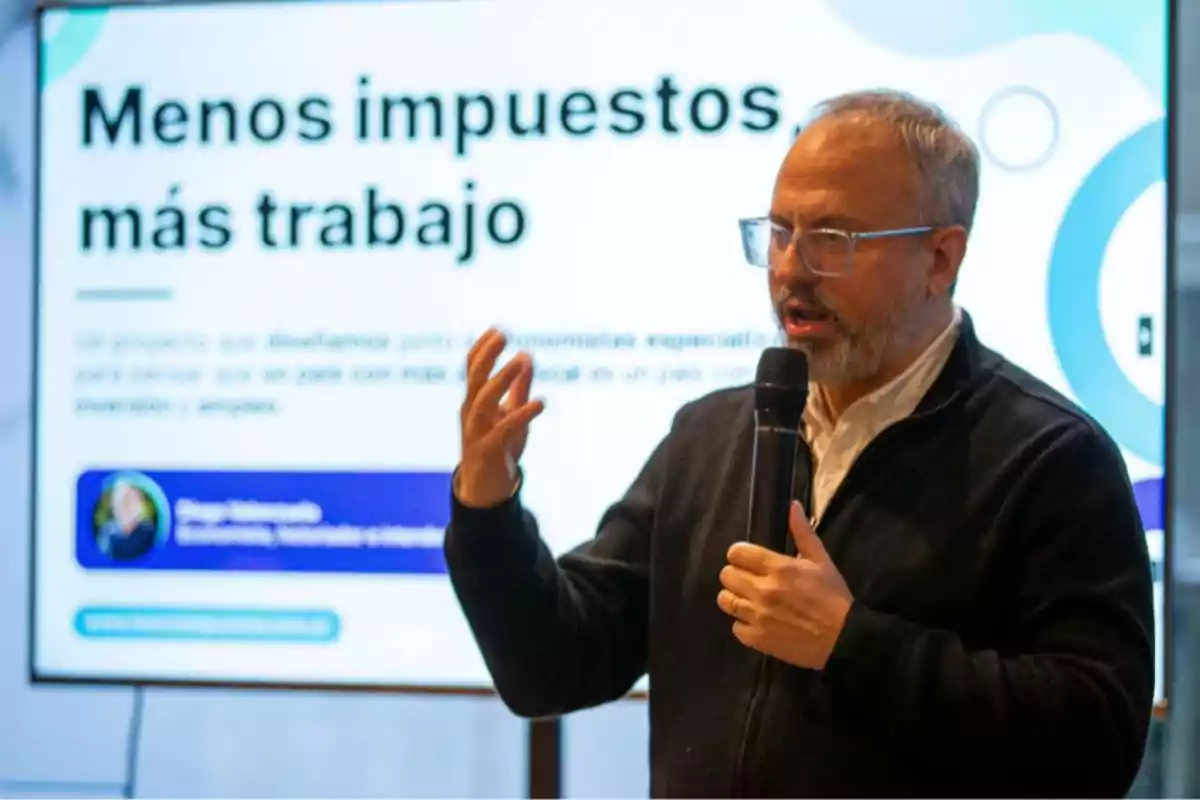 Hombre de mediana edad con gafas y barba hablando por micrófono frente a una pantalla que muestra el texto Menos impuestos más trabajo