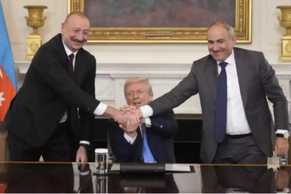 Three men in suits smile as they shake hands in front of a table in an elegant room. Three men in suits smile as they shake hands in front of a table in an elegant room.