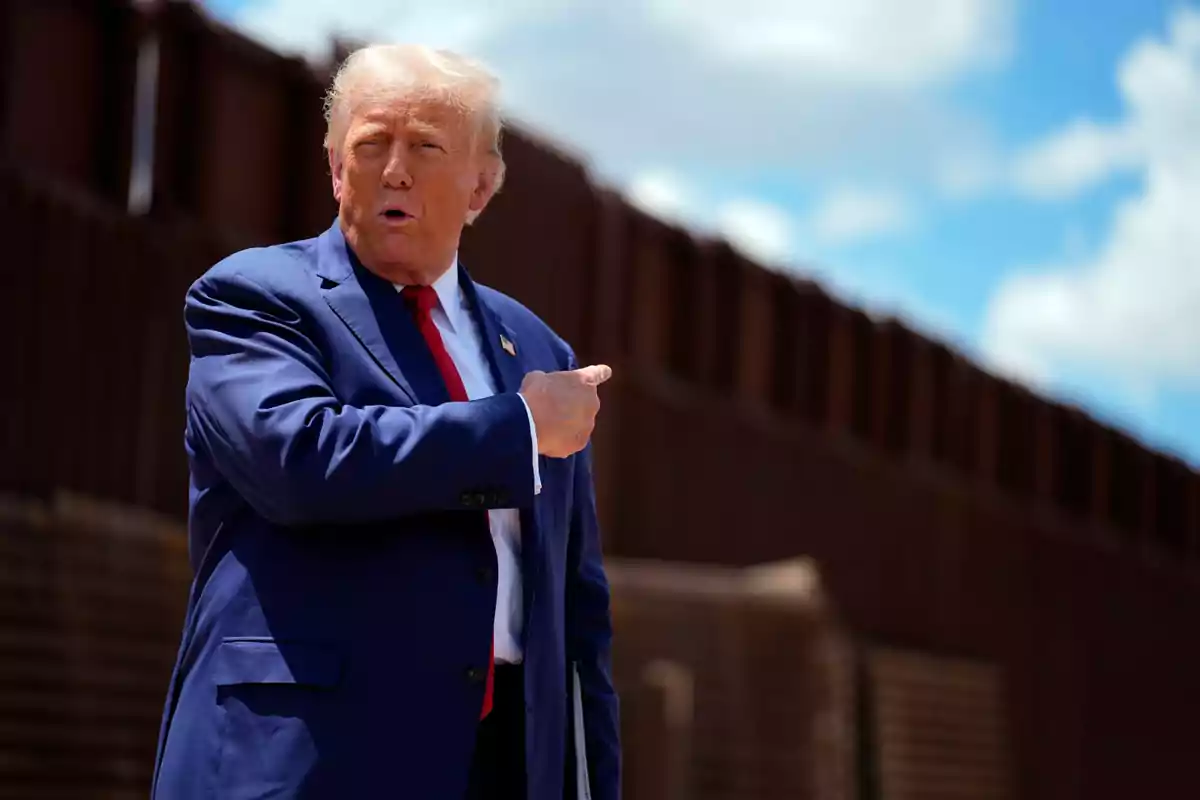 Blond-haired man in a blue suit and red tie pointing to the side in front of a metal fence under a partly cloudy sky