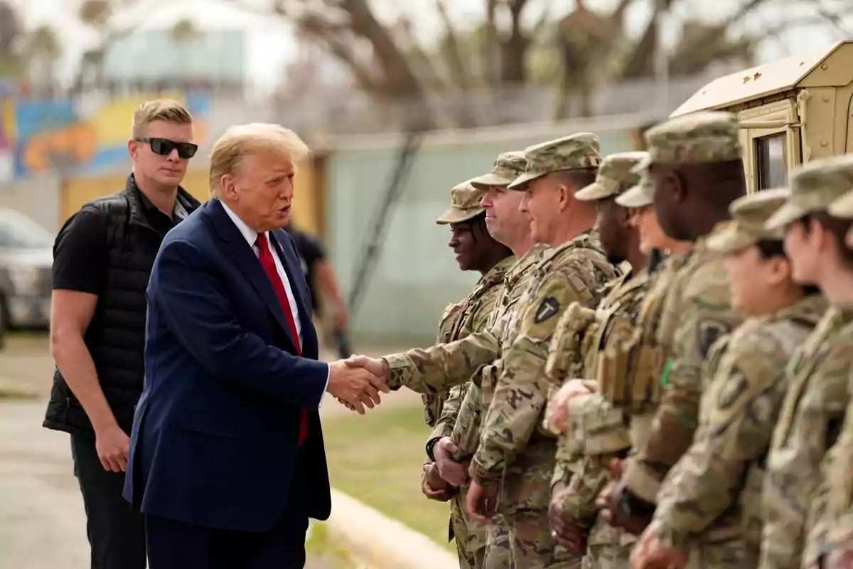 A man in a blue suit and red tie greets a group of uniformed soldiers standing in line while another person wearing sunglasses watches in the background. A man in a blue suit and red tie greets a group of uniformed soldiers standing in line while another person wearing sunglasses watches in the background.