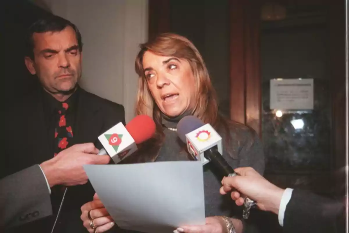 A woman holds a sheet of paper while giving statements to the press surrounded by microphones, and a man watches her closely. A woman holds a sheet of paper while giving statements to the press surrounded by microphones, and a man watches her closely.