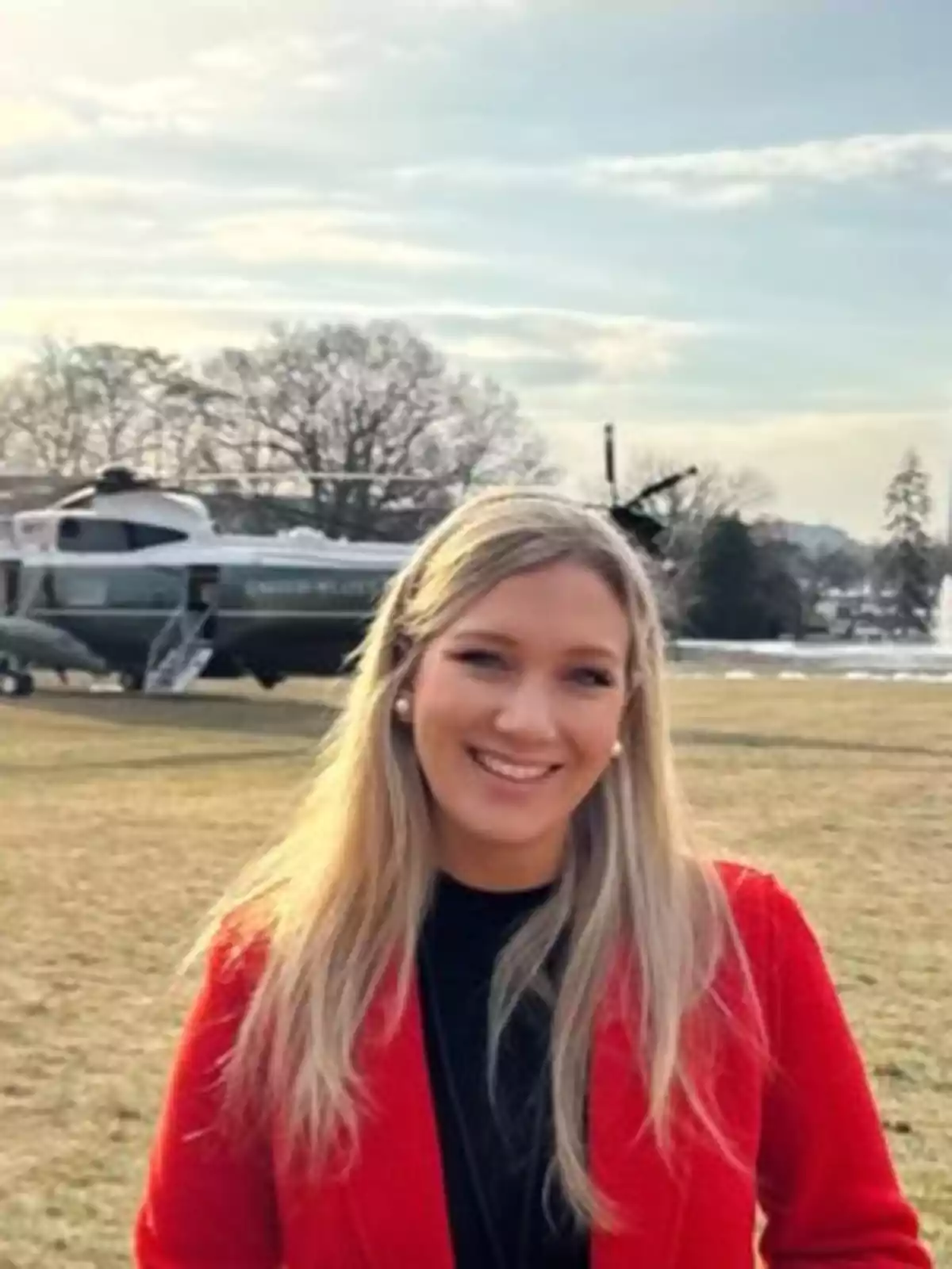 Blonde woman in a red coat smiling outdoors with a helicopter and trees in the background Blonde woman in a red coat smiling outdoors with a helicopter and trees in the background