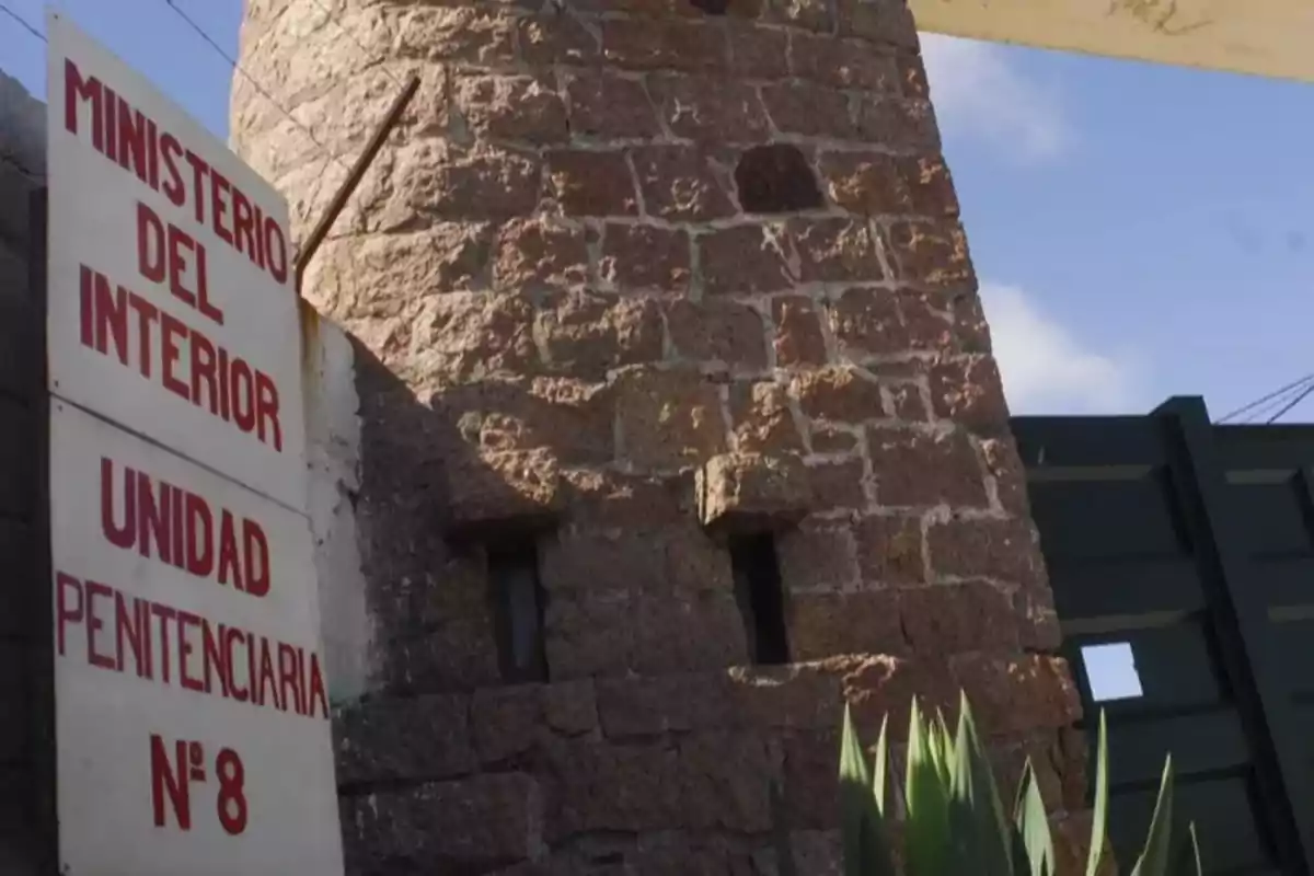 Cartel con letras rojas que dice Ministerio del Interior Unidad Penitenciaria N° 8 junto a una torre de piedra y una planta en primer plano