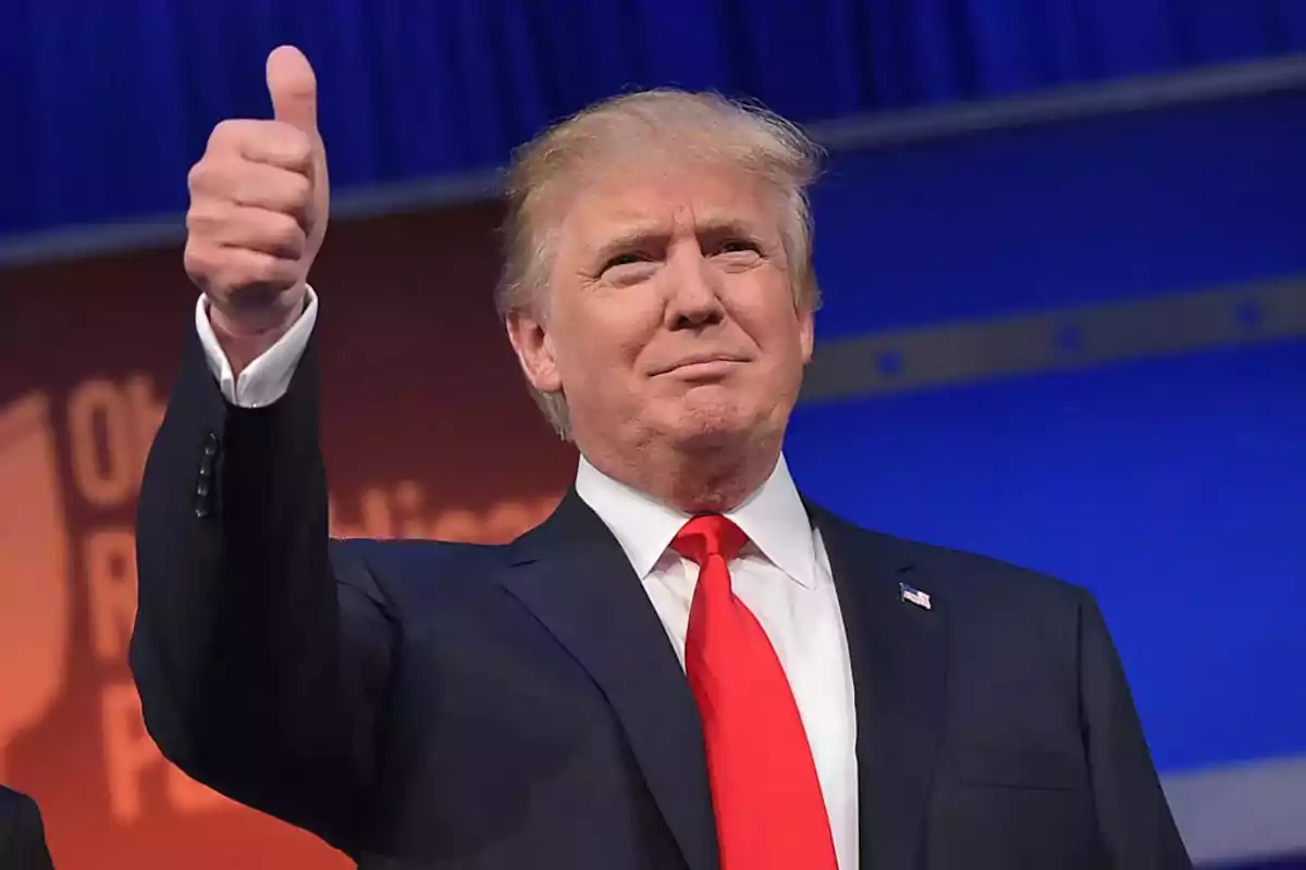 Blond-haired man in a dark suit and red tie giving a thumbs up in approval