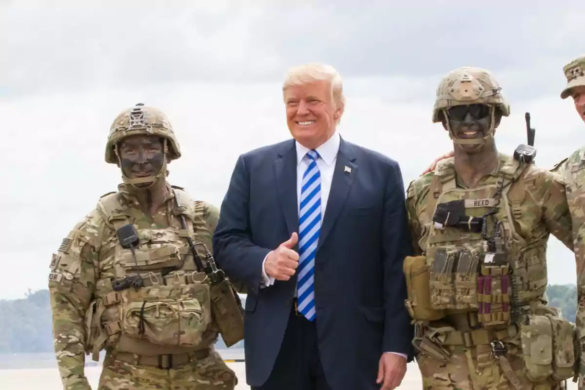 Man in a suit and blue tie posing and smiling next to two soldiers in camouflage uniforms and military gear Man in a suit and blue tie posing and smiling next to two soldiers in camouflage uniforms and military gear