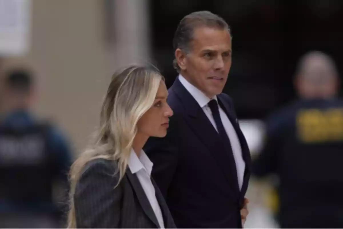 Two people dressed in suits walk together outdoors with blurred people in the background.
