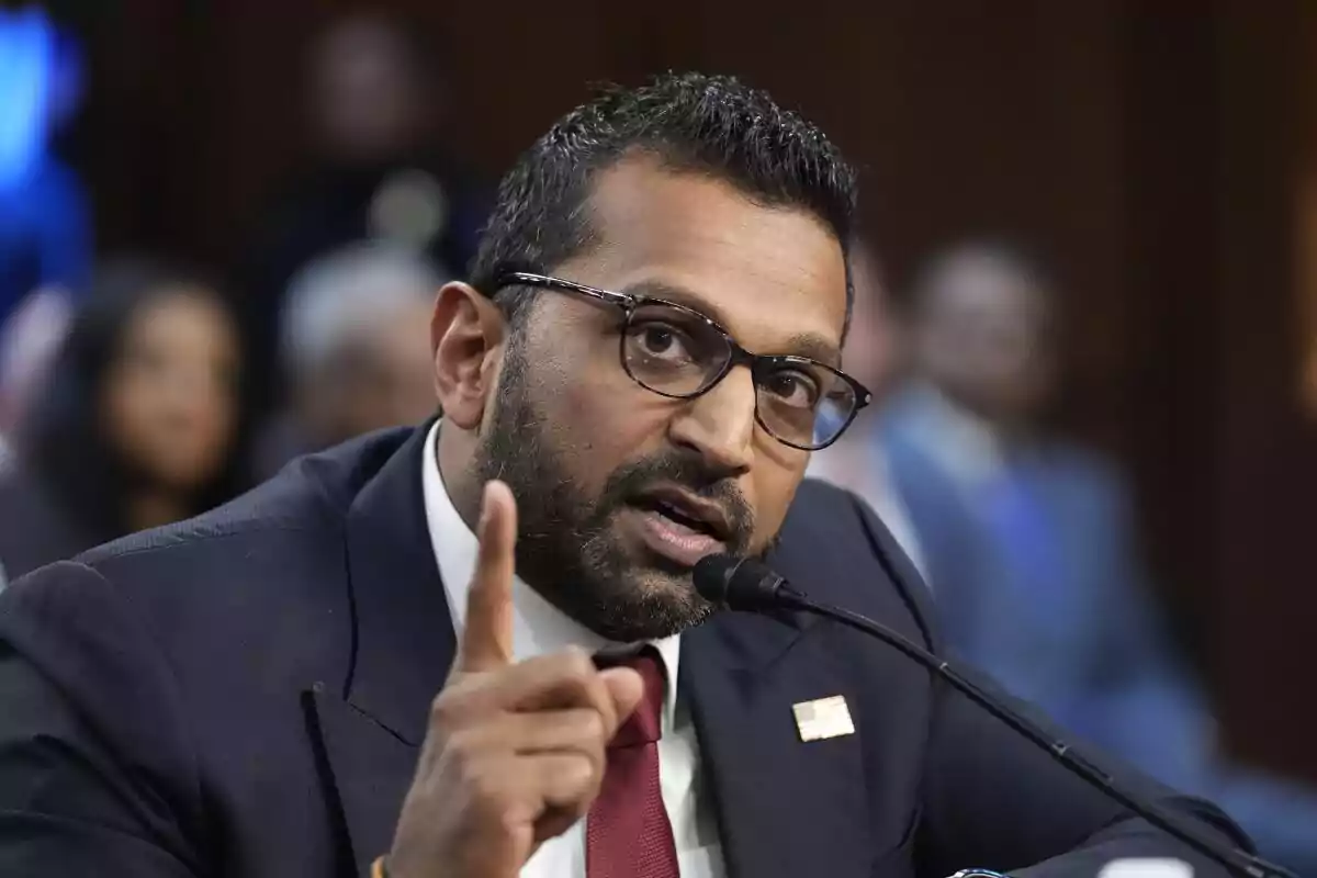 Man with glasses and a beard speaking into a microphone while raising his index finger for emphasis