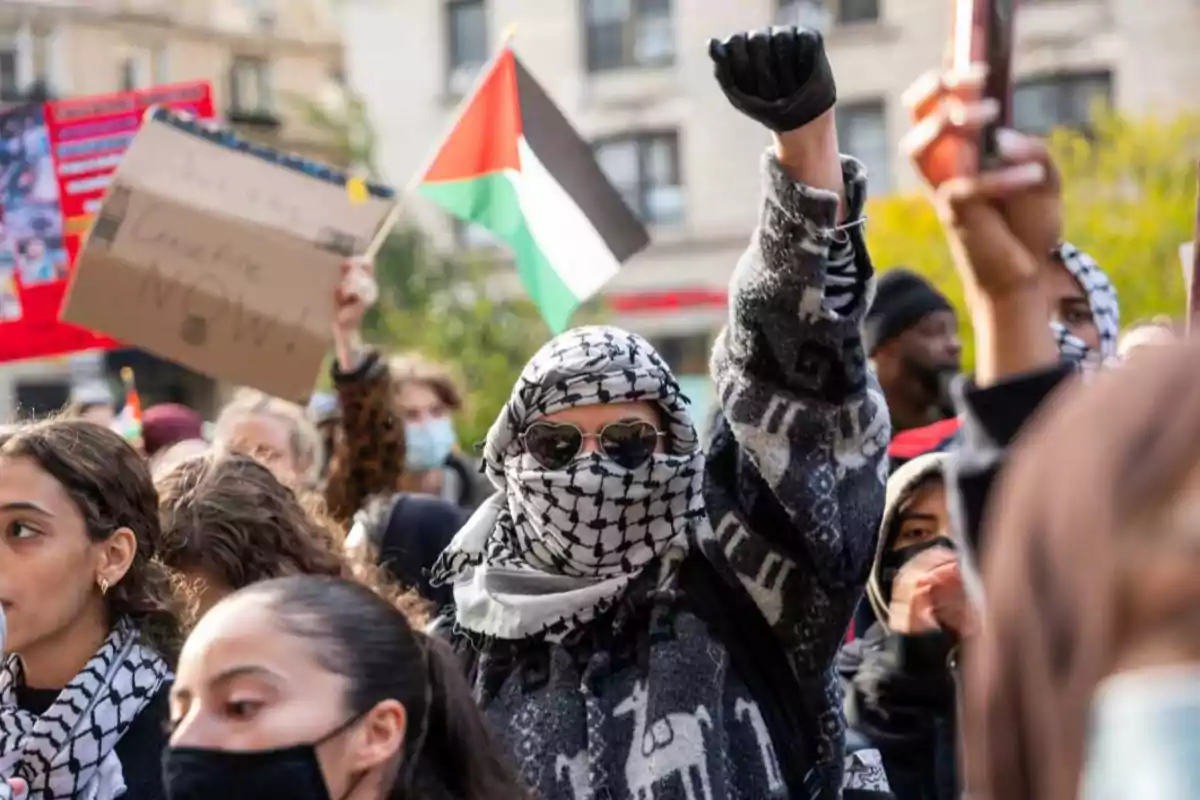 Personas manifestándose con pañuelos palestinos, una bandera de Palestina y carteles en un ambiente urbano Personas manifestándose con pañuelos palestinos, una bandera de Palestina y carteles en un ambiente urbano