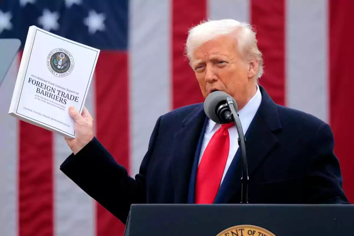Hombre de cabello rubio con traje oscuro y corbata roja sostiene un libro titulado Foreign Trade Barriers mientras habla frente a un micrófono con una bandera de Estados Unidos de fondo