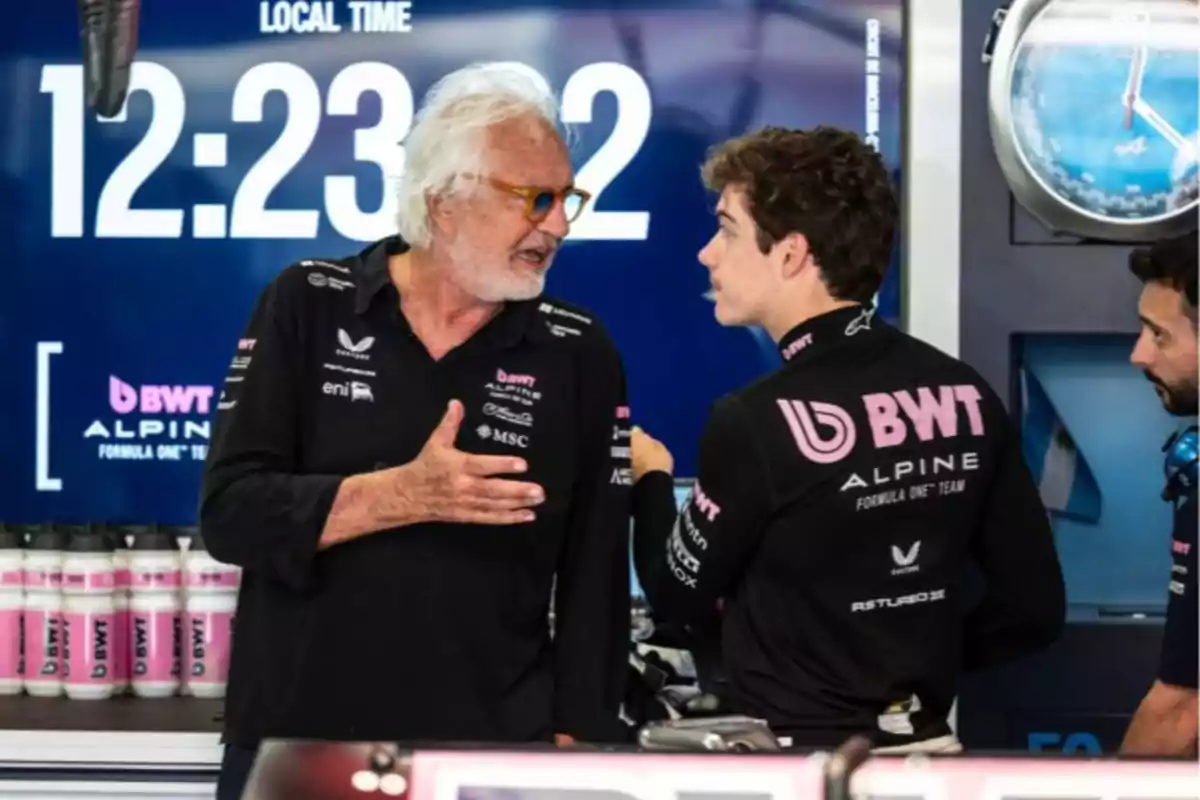 Two members of the Alpine Formula 1 team are talking in the garage, with a large digital clock in the background and pink bottles lined up on a table.