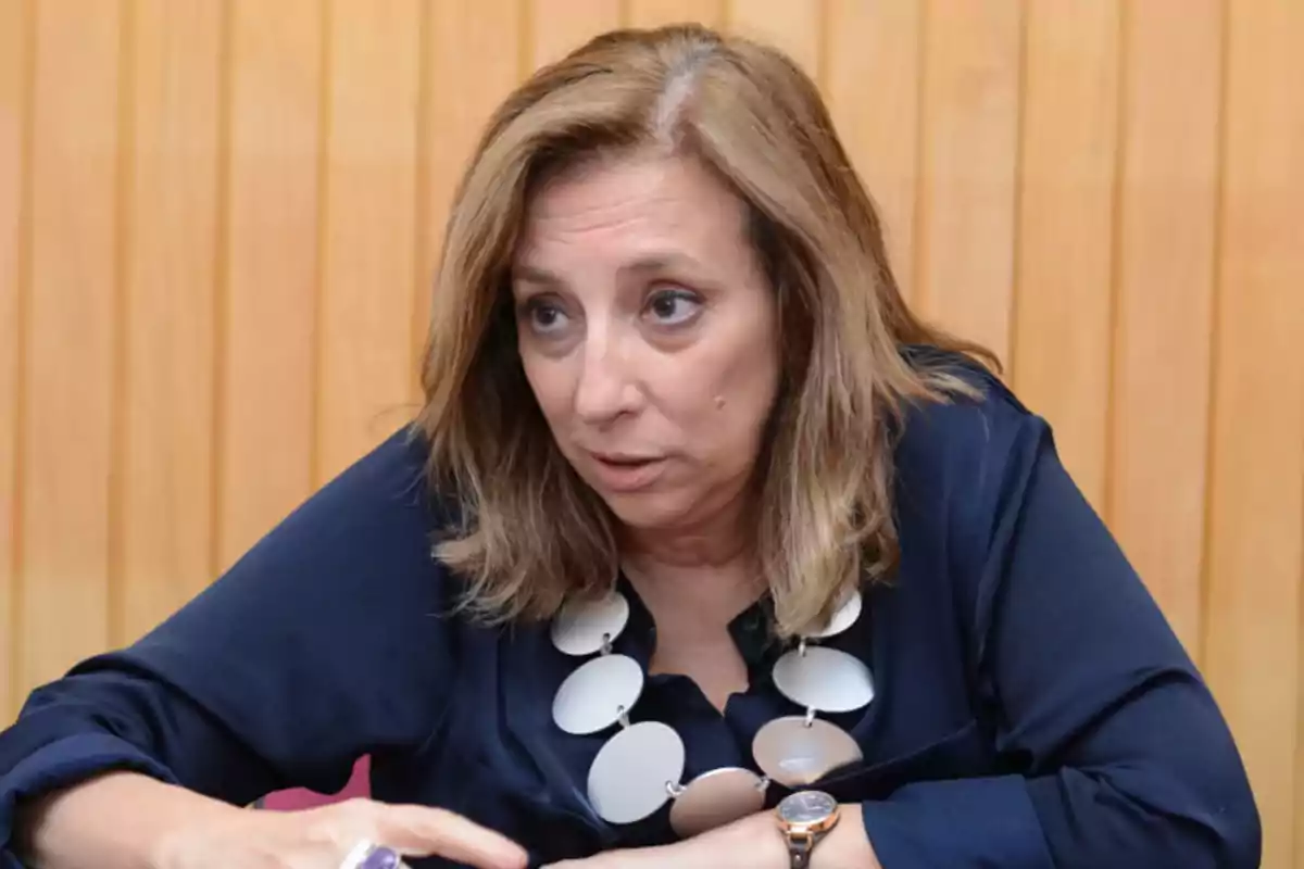 Mujer de cabello castaño claro con blusa azul oscuro y collar de grandes eslabones circulares sentada frente a una pared de madera Mujer de cabello castaño claro con blusa azul oscuro y collar de grandes eslabones circulares sentada frente a una pared de madera