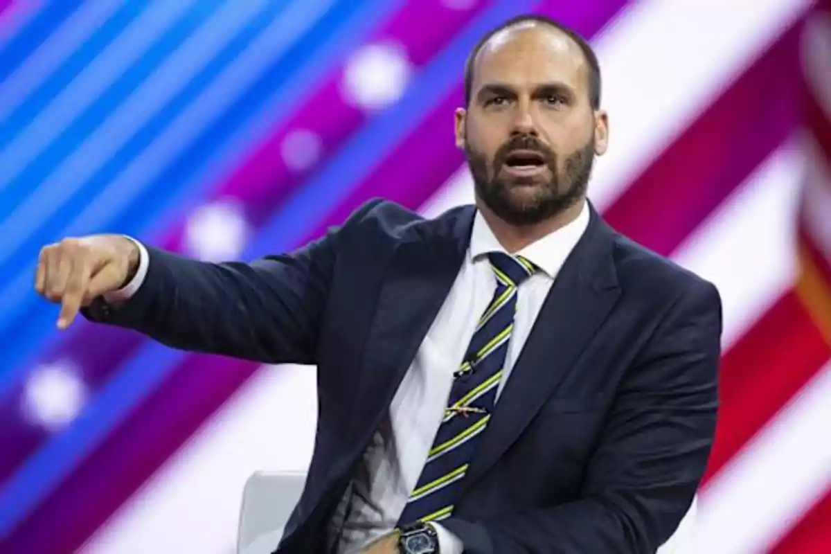 Bearded man in a dark suit speaking and gesturing with his arm extended against a colorful background with diagonal lines