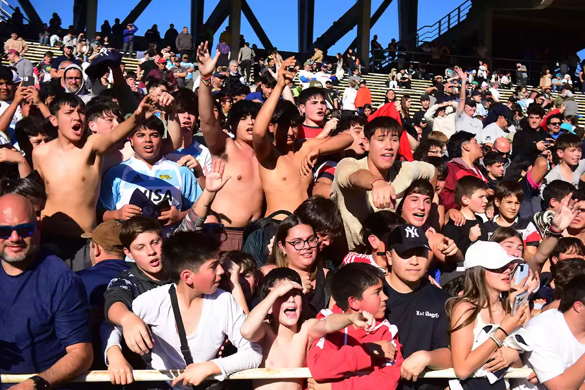 A large group of enthusiastic young people and children in the stands of a stadium enjoying a sporting event under the sun.