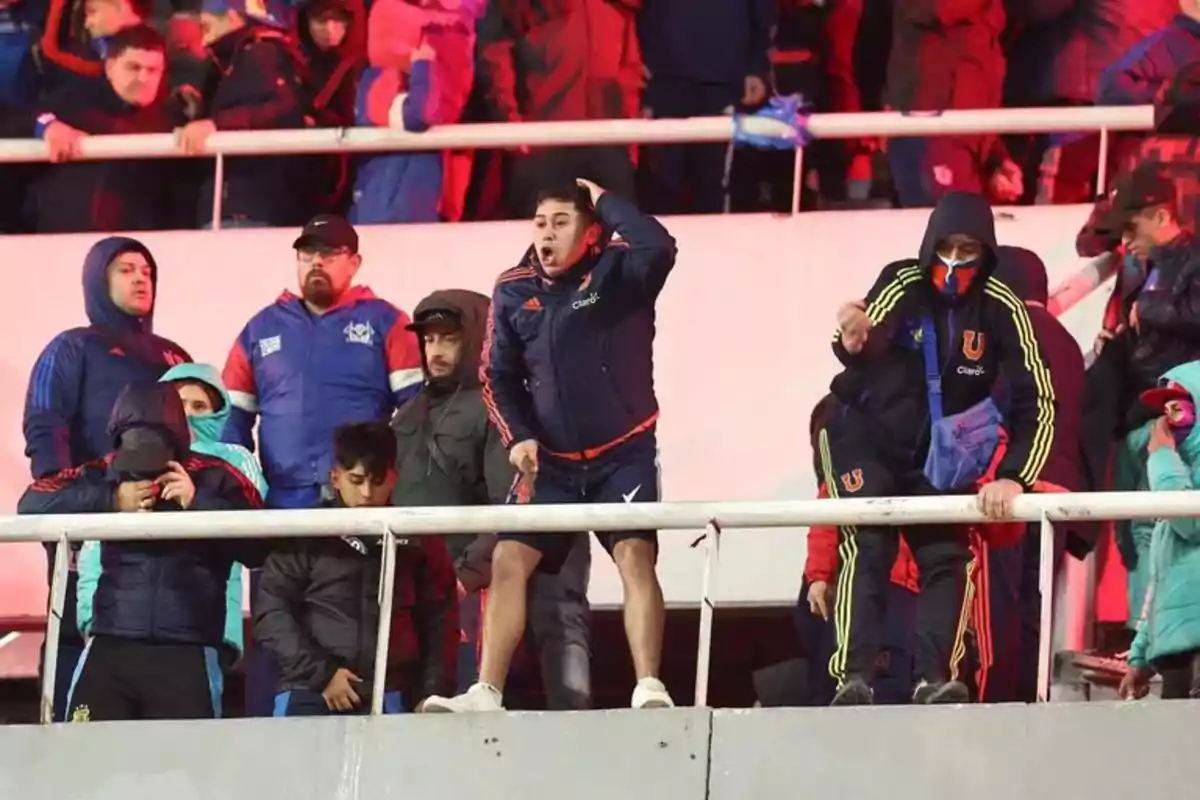 Soccer fans wearing winter clothing and University of Chile sweatjerseys display various emotions in the stands of a stadium lit with red lights. Soccer fans wearing winter clothing and University of Chile sweatjerseys display various emotions in the stands of a stadium lit with red lights.