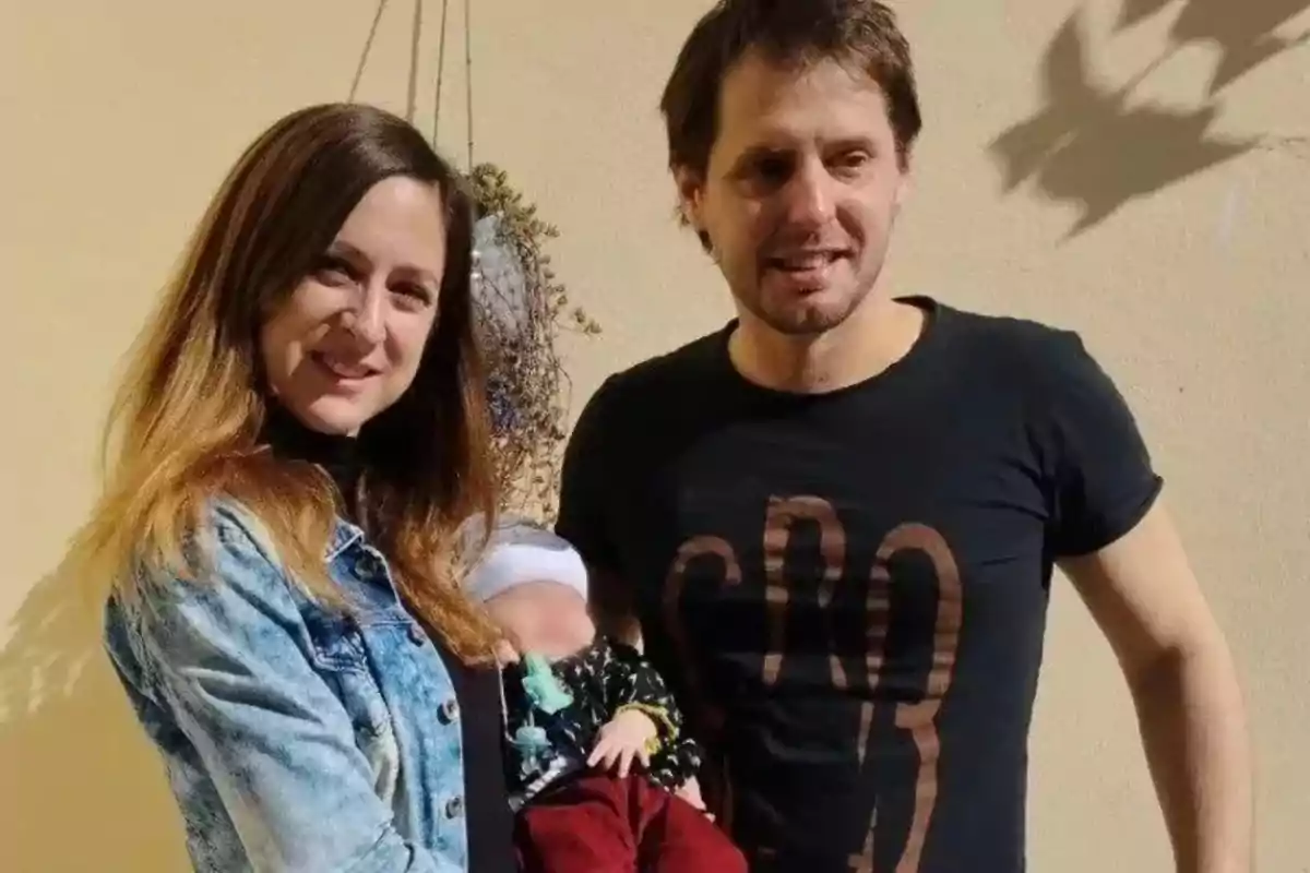 A smiling couple poses outdoors with a baby in their arms, with a light-colored wall in the background and natural light.