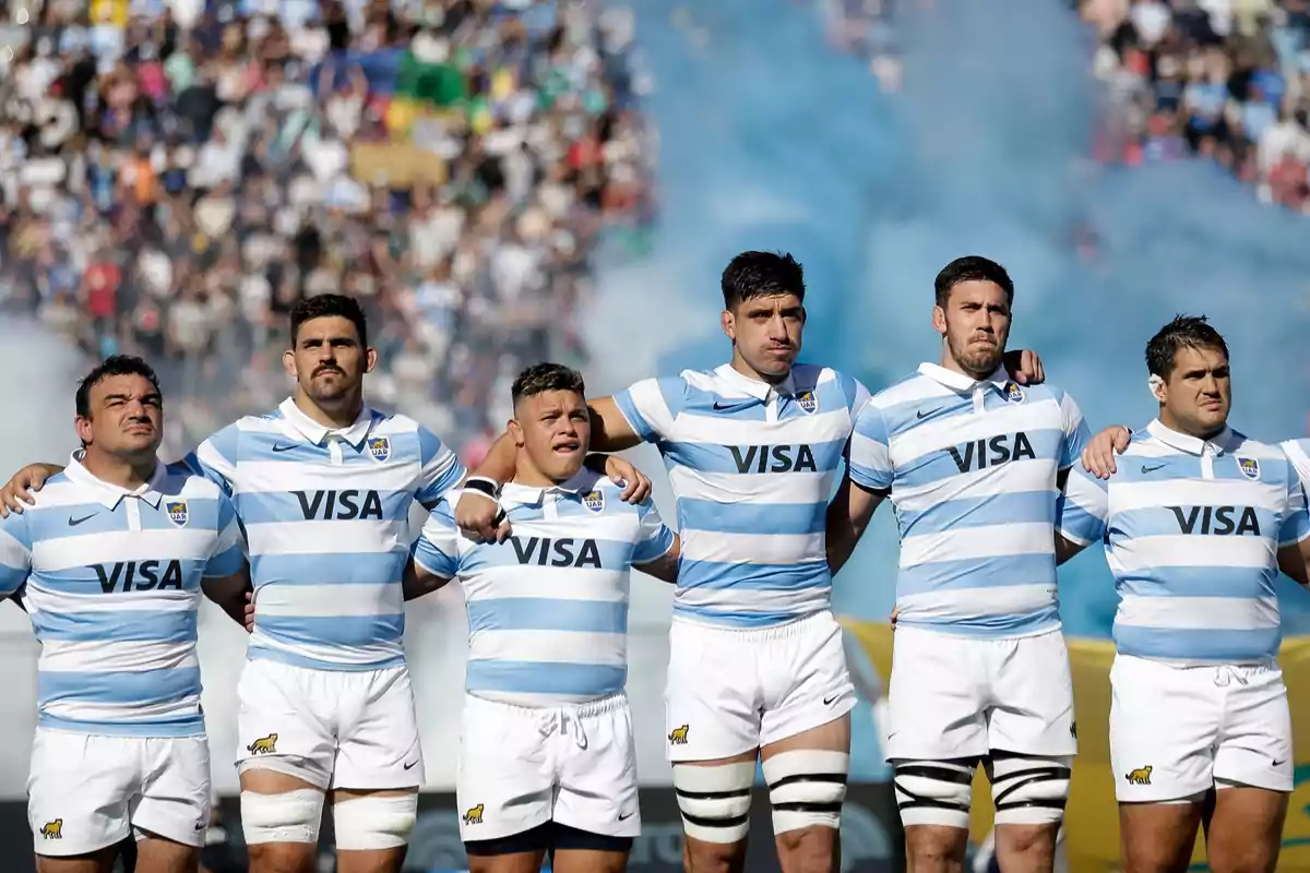 Argentinian rugby team players lined up with their arms linked before a match, wearing light blue and white jerseys with the Visa logo, with blue smoke and a crowd in the background.