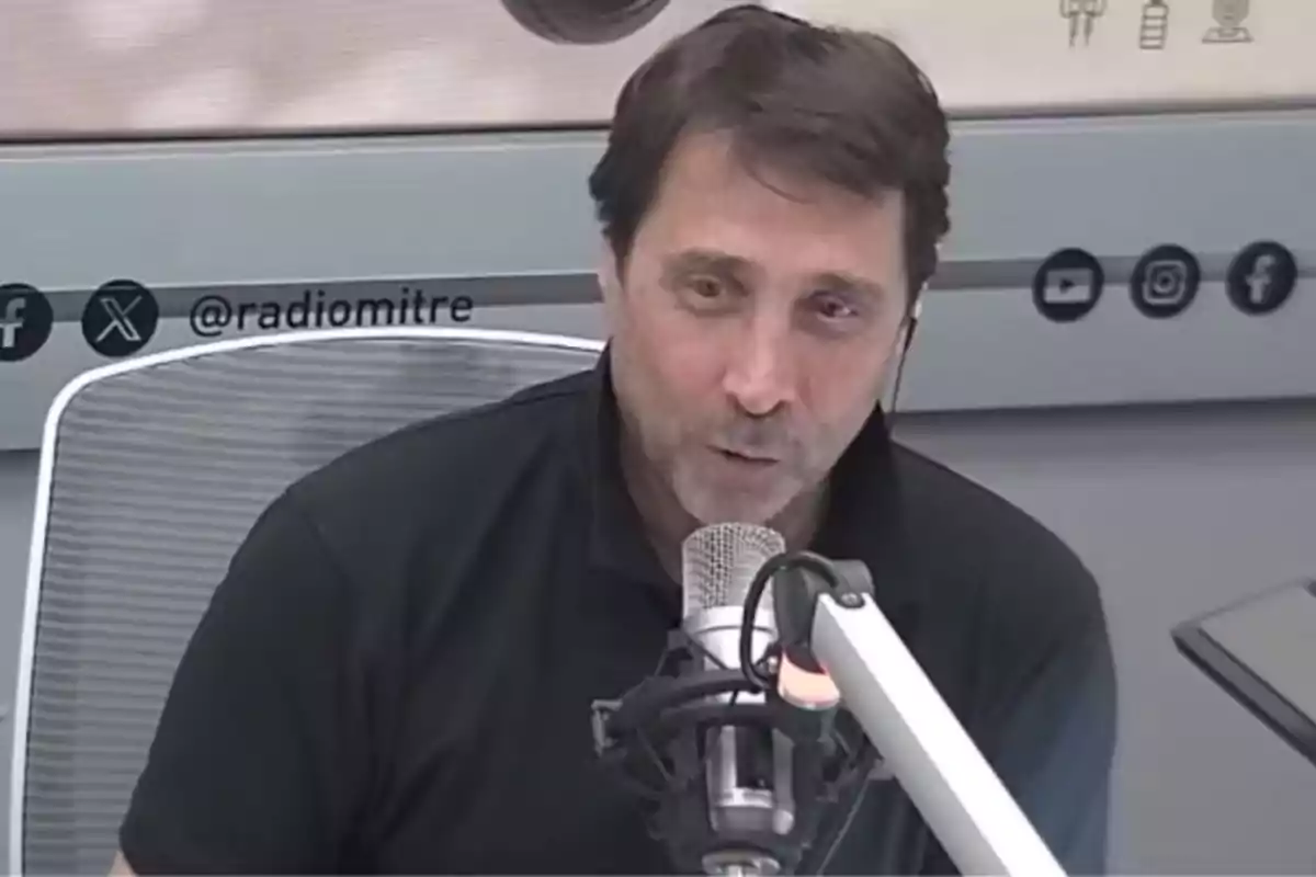 Man speaking in front of a microphone in a radio studio with the social media user @radiomitre visible in the background Man speaking in front of a microphone in a radio studio with the social media user @radiomitre visible in the background
