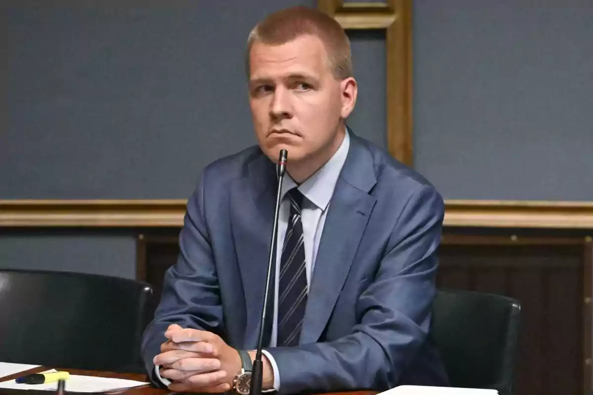 Short-haired blond man in a blue suit sitting at a table in front of a microphone with a serious expression Short-haired blond man in a blue suit sitting at a table in front of a microphone with a serious expression