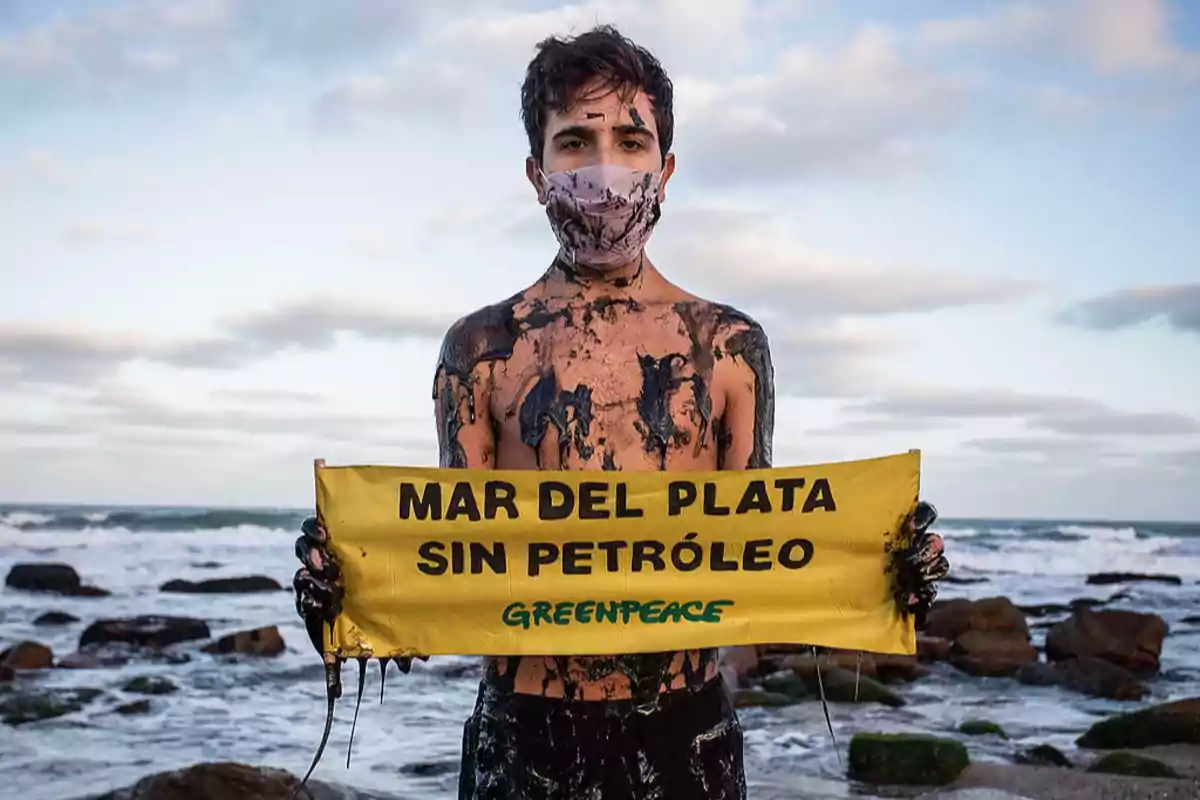 Young man covered in oil holds a yellow sign with the message “Mar del Plata without oil” from Greenpeace on a rocky beach.