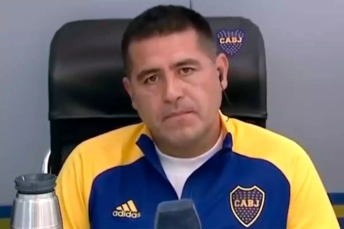Hombre con chaqueta de Boca Juniors sentado frente a un micrófono y un termo
