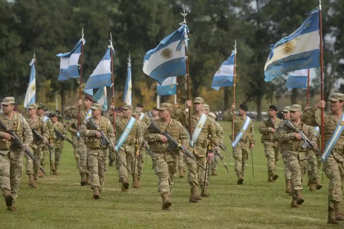 Soldiers marching in formation on grass, wearing camouflage uniforms, carrying rifles and Argentine flags, in an outdoor setting with trees in the background. Soldiers marching in formation on grass, wearing camouflage uniforms, carrying rifles and Argentine flags, in an outdoor setting with trees in the background.