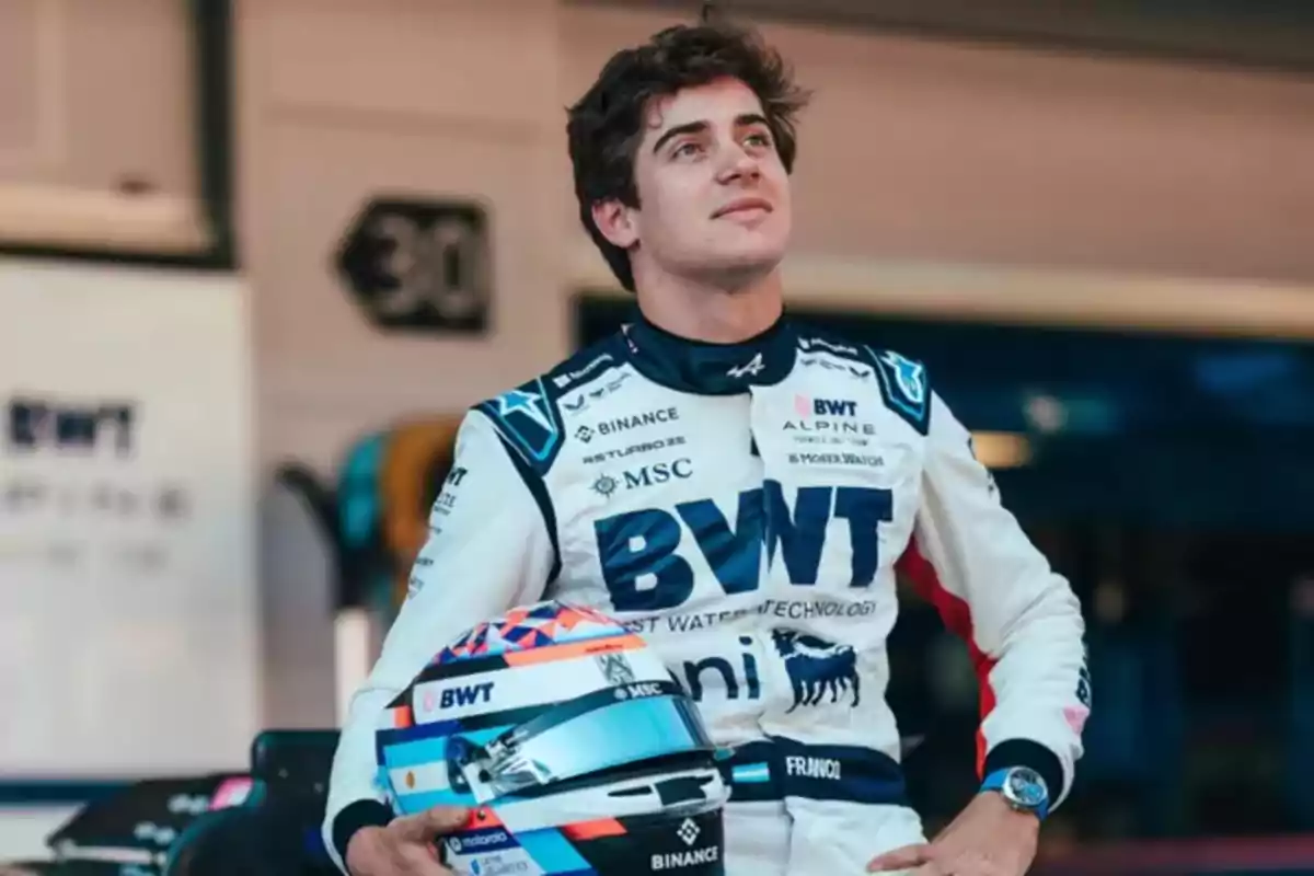 Auto racing driver in a white and blue racing suit holding a helmet in a motorsport team garage