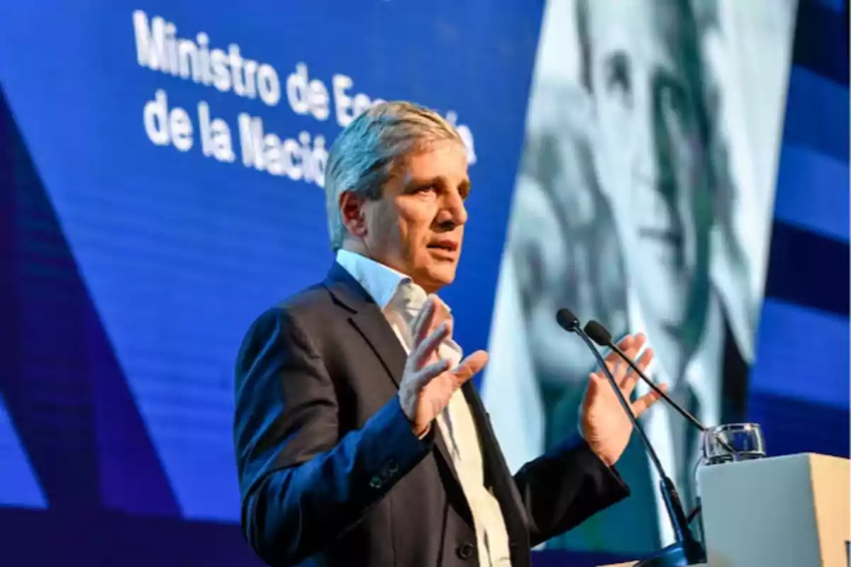 Hombre de cabello canoso hablando en un podio con micrófonos frente a una pantalla azul que muestra texto y una imagen en blanco y negro de fondo